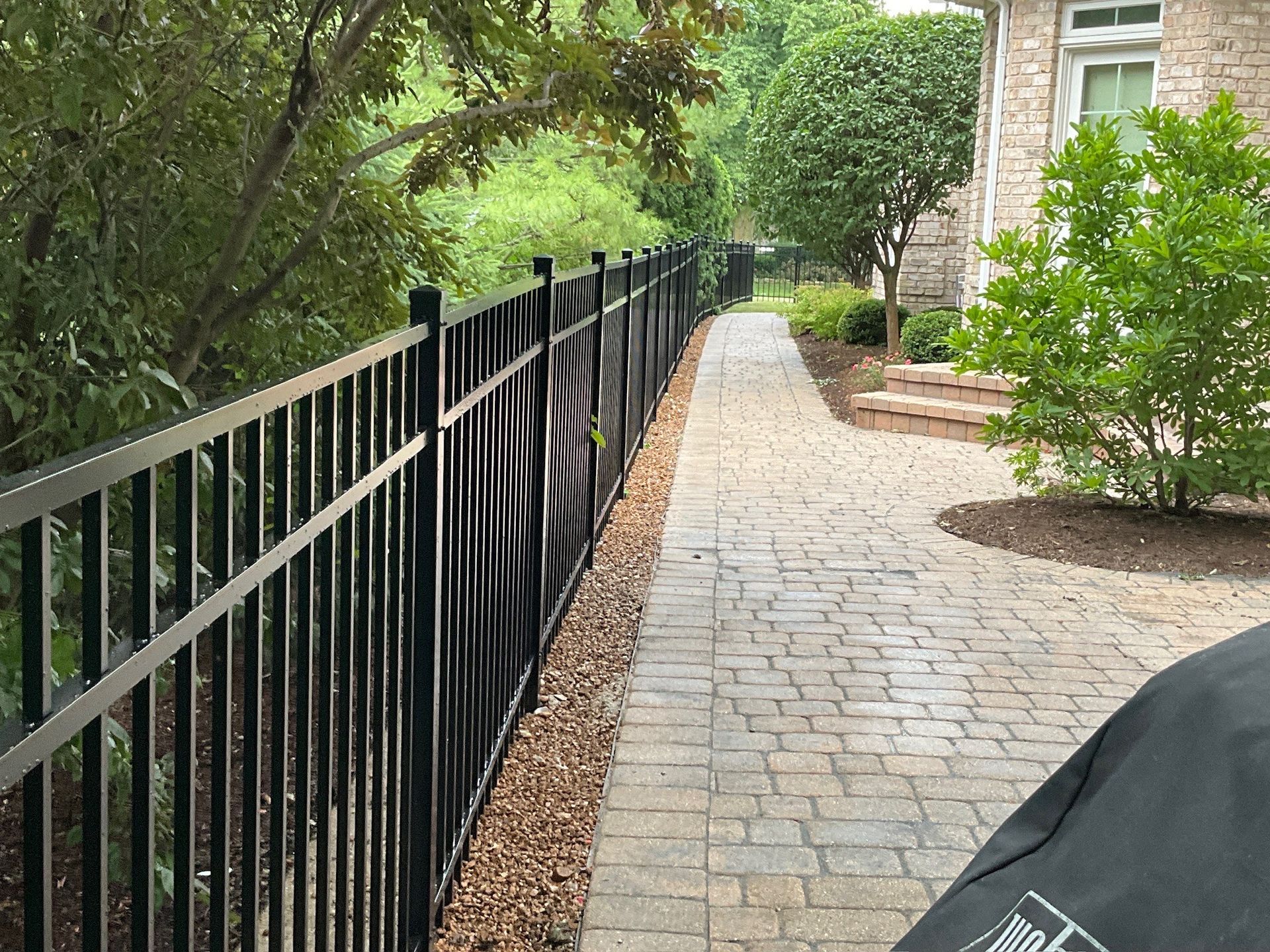 Black metal fence along a stone walkway bordered by landscaping and a house.