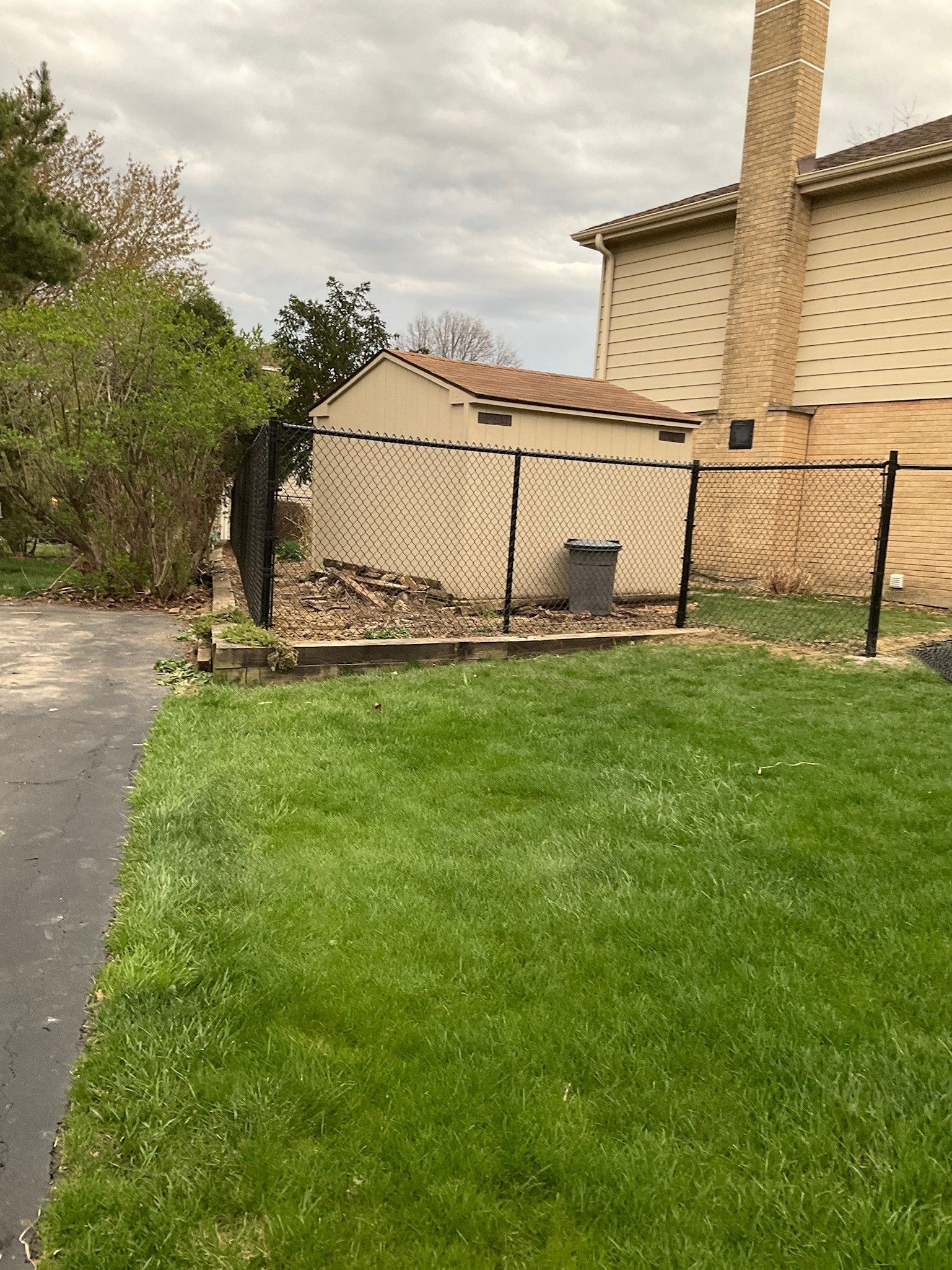 Green lawn, asphalt path, chain-link fence, beige shed, and a house with a brick chimney under a cloudy sky.