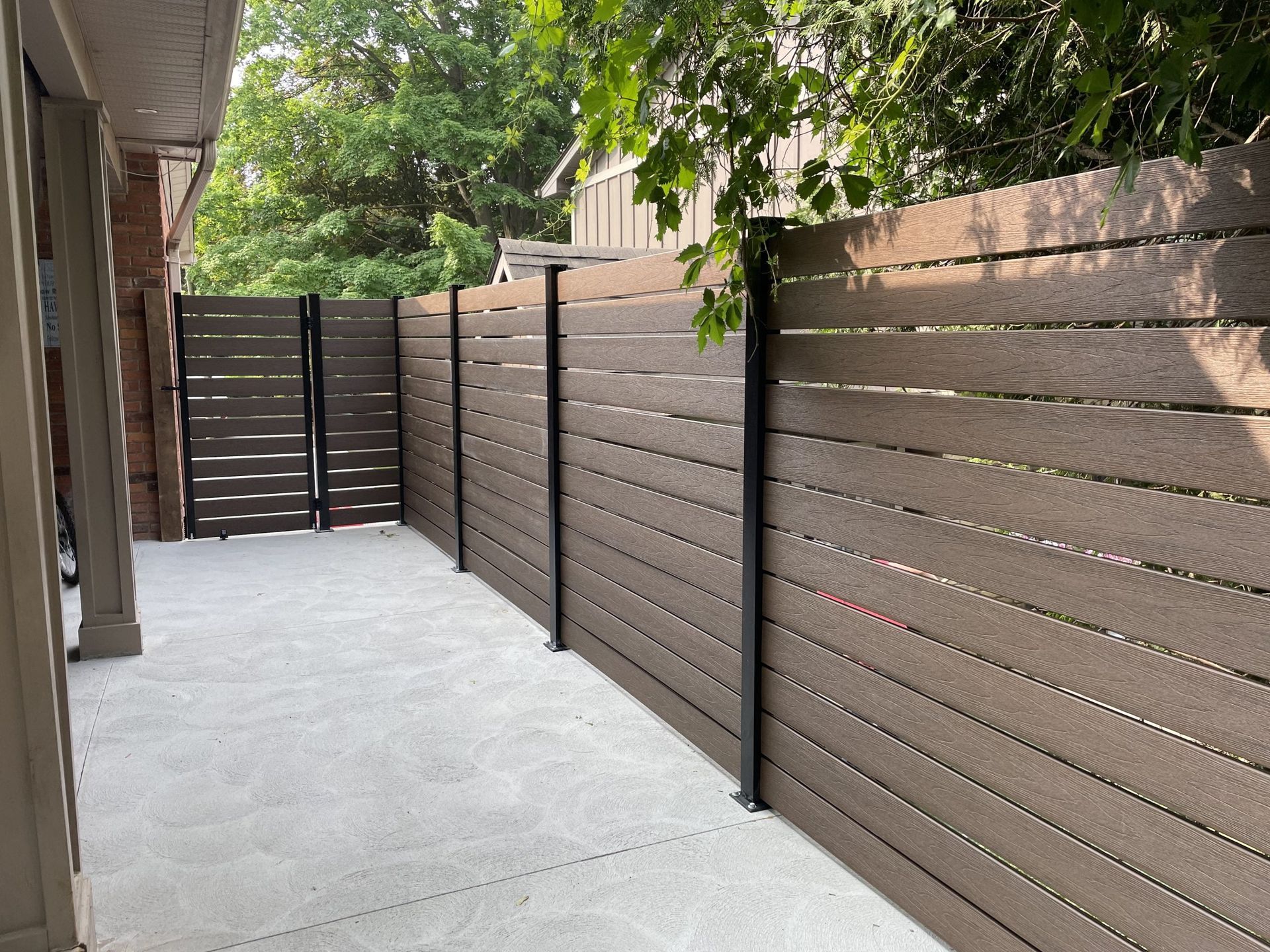 Brown horizontal slat fence with black posts along a concrete walkway, next to a building.