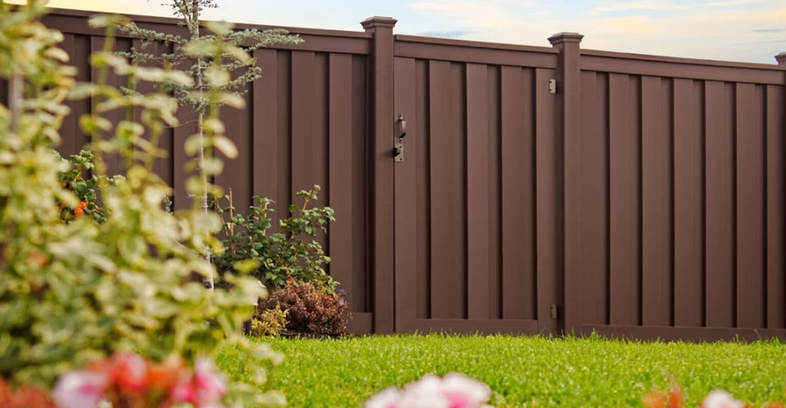Brown vinyl fence with gate in a grassy yard, surrounded by green shrubs and a blurred foreground of flowers.