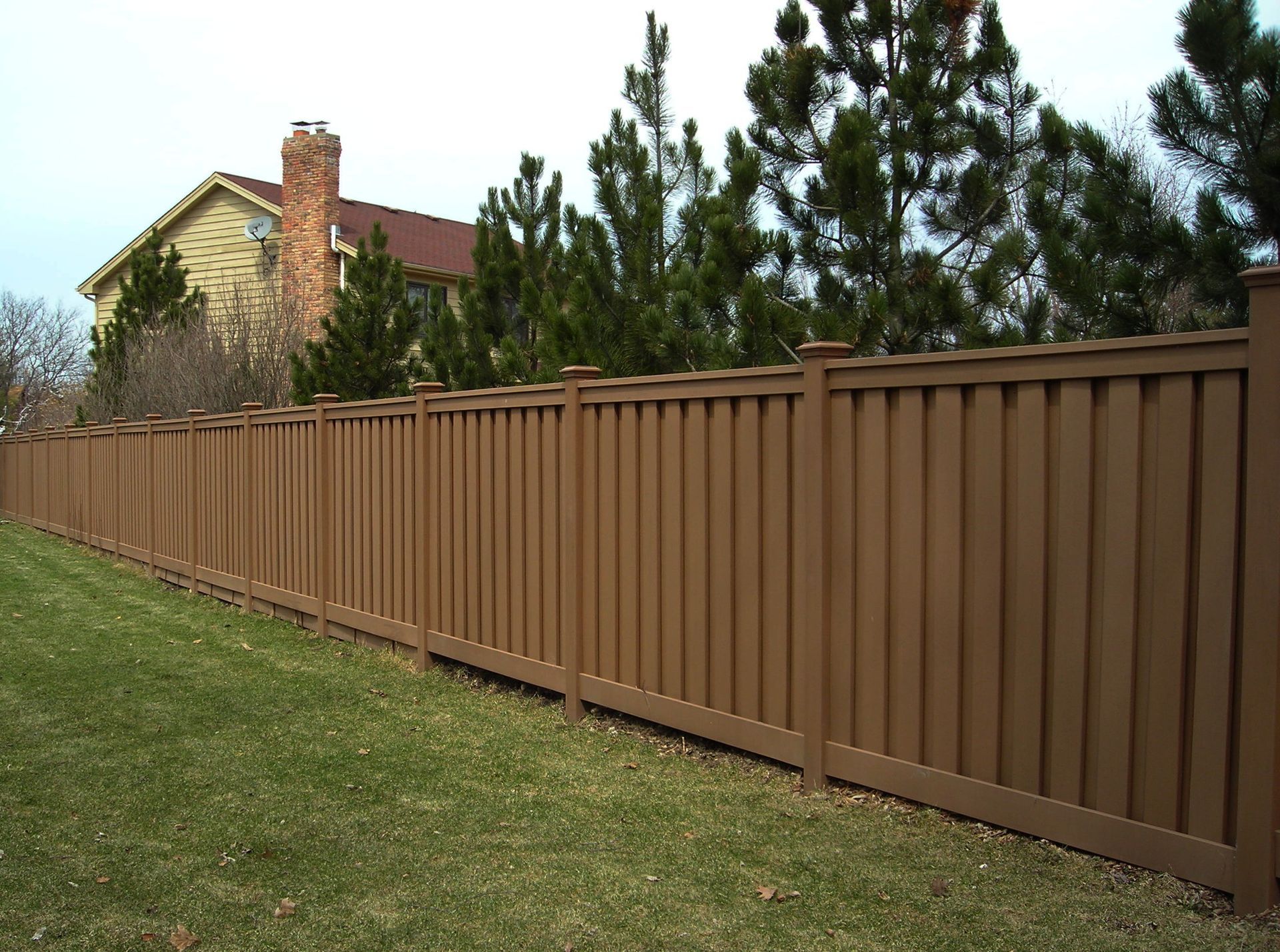 Brown vinyl fence bordering green lawn, with house and trees in the background.