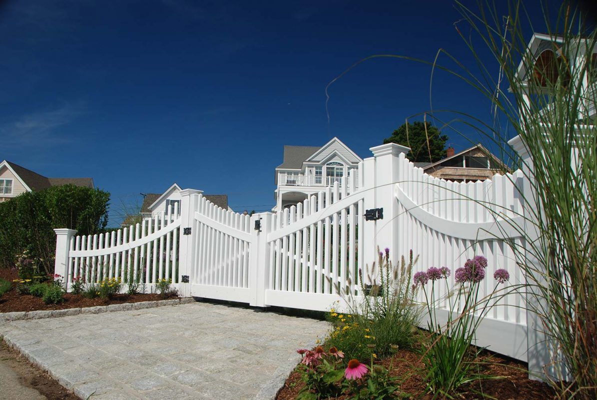 White picket fence with gate in front of houses; gravel driveway, blue sky.