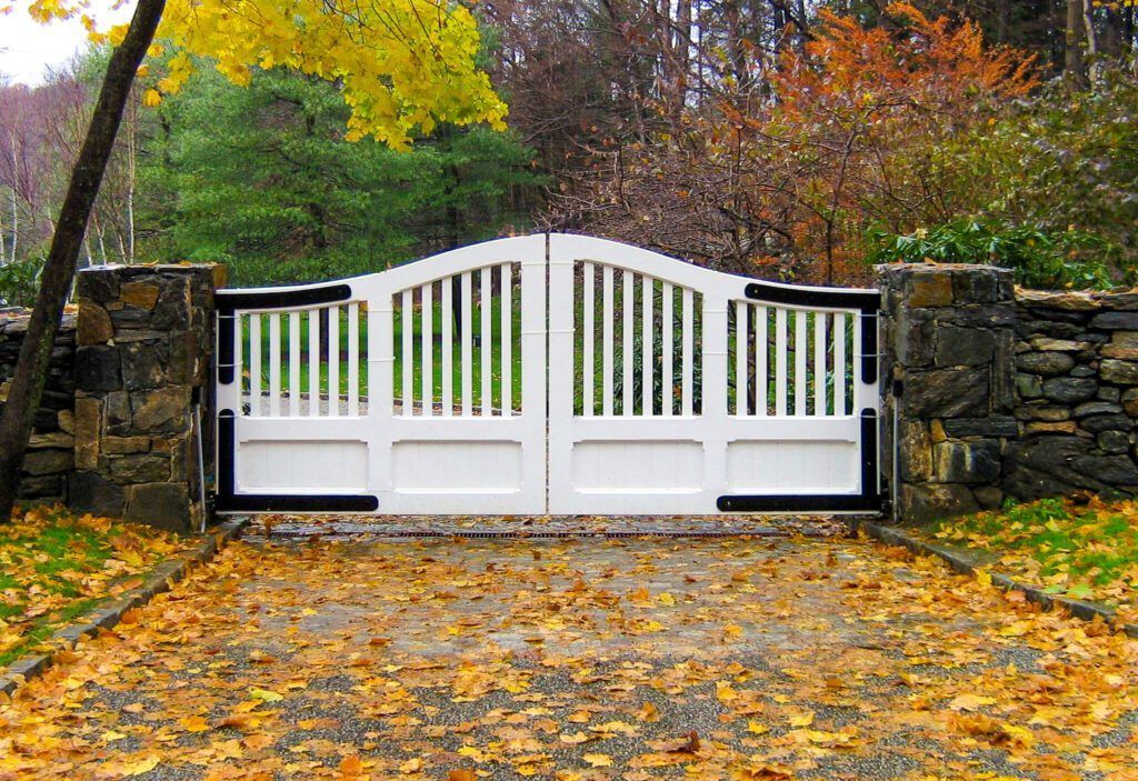 White gate on stone pillars, autumn leaves on the driveway, trees in background.