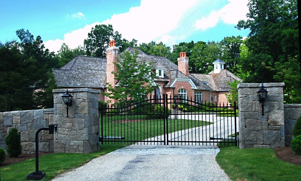 A large brick house behind a black gate. Stone pillars flank the driveway entrance.