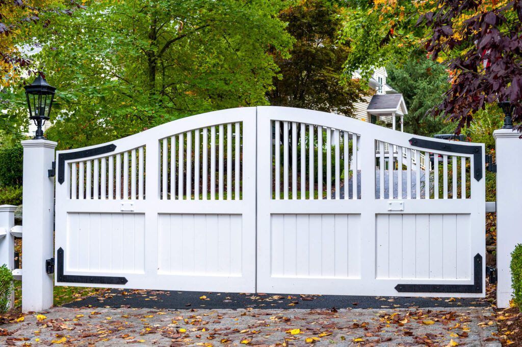 White wooden driveway gate with black hinges and vertical bars, autumn leaves on the ground.