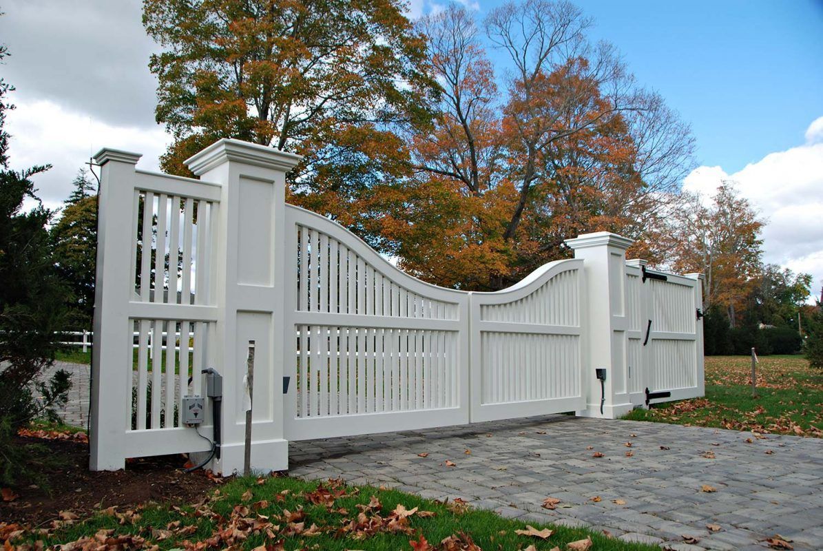White wooden driveway gate, arched design, brick pavers, autumn leaves, trees in background.