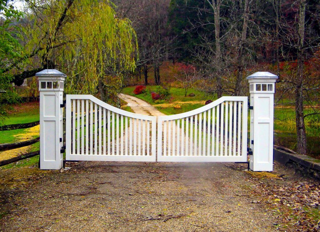 White picket gates on gravel driveway, flanked by white pillars, leading to path with foliage.