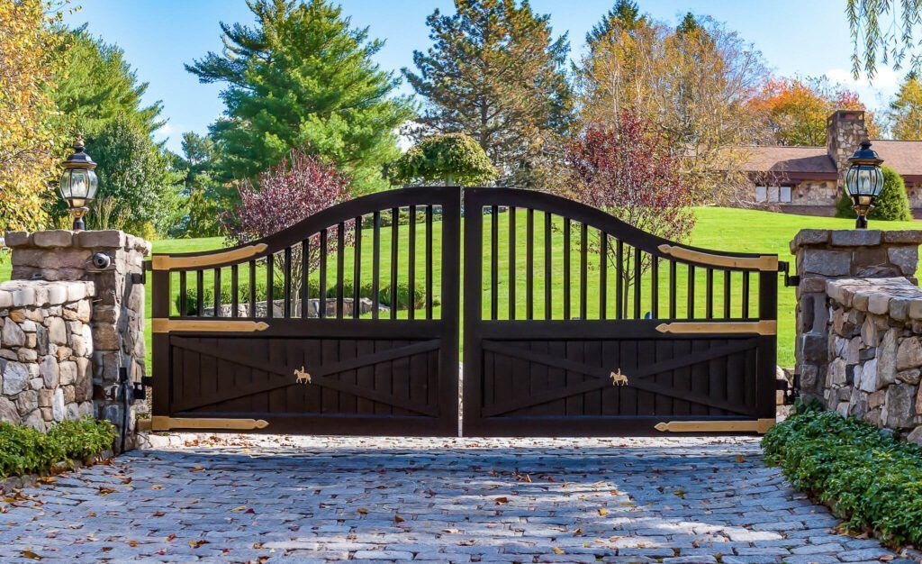Dark wood driveway gates open on a brick driveway, with stone pillars, and green lawn with trees.