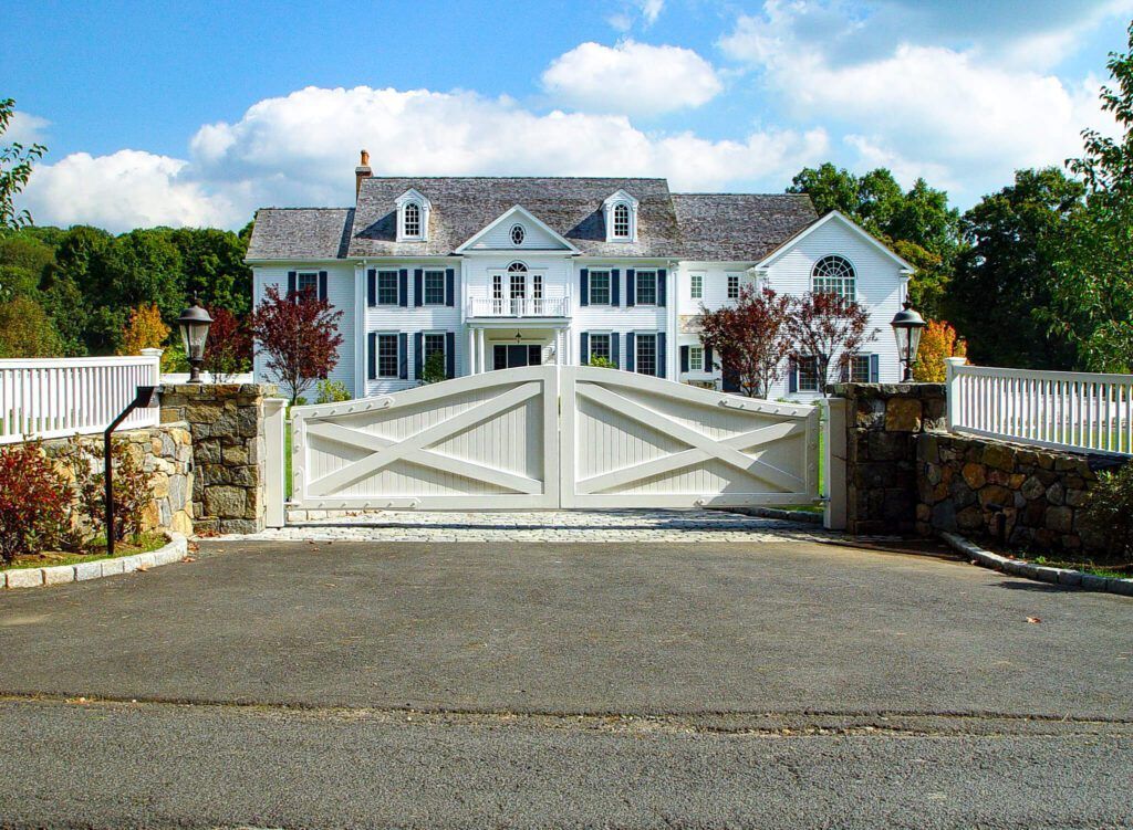 White mansion with gray roof behind white gate, set in a tree-lined landscape under a cloudy blue sky.