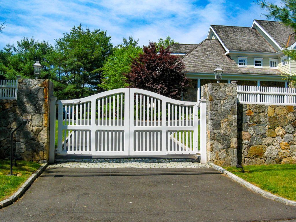 White gated entrance to a house with stone pillars, asphalt driveway, and green lawn.