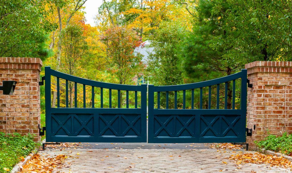 Blue gated entrance with brick pillars, leading to a driveway, framed by fall foliage.