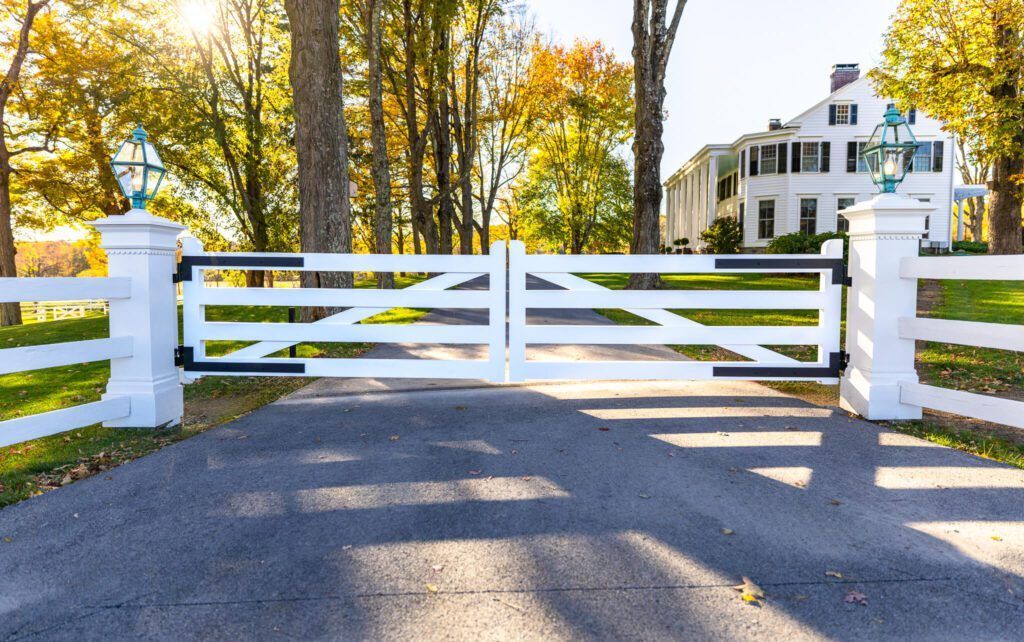 White gate and fence leading to a large white house on a sunny day with trees.