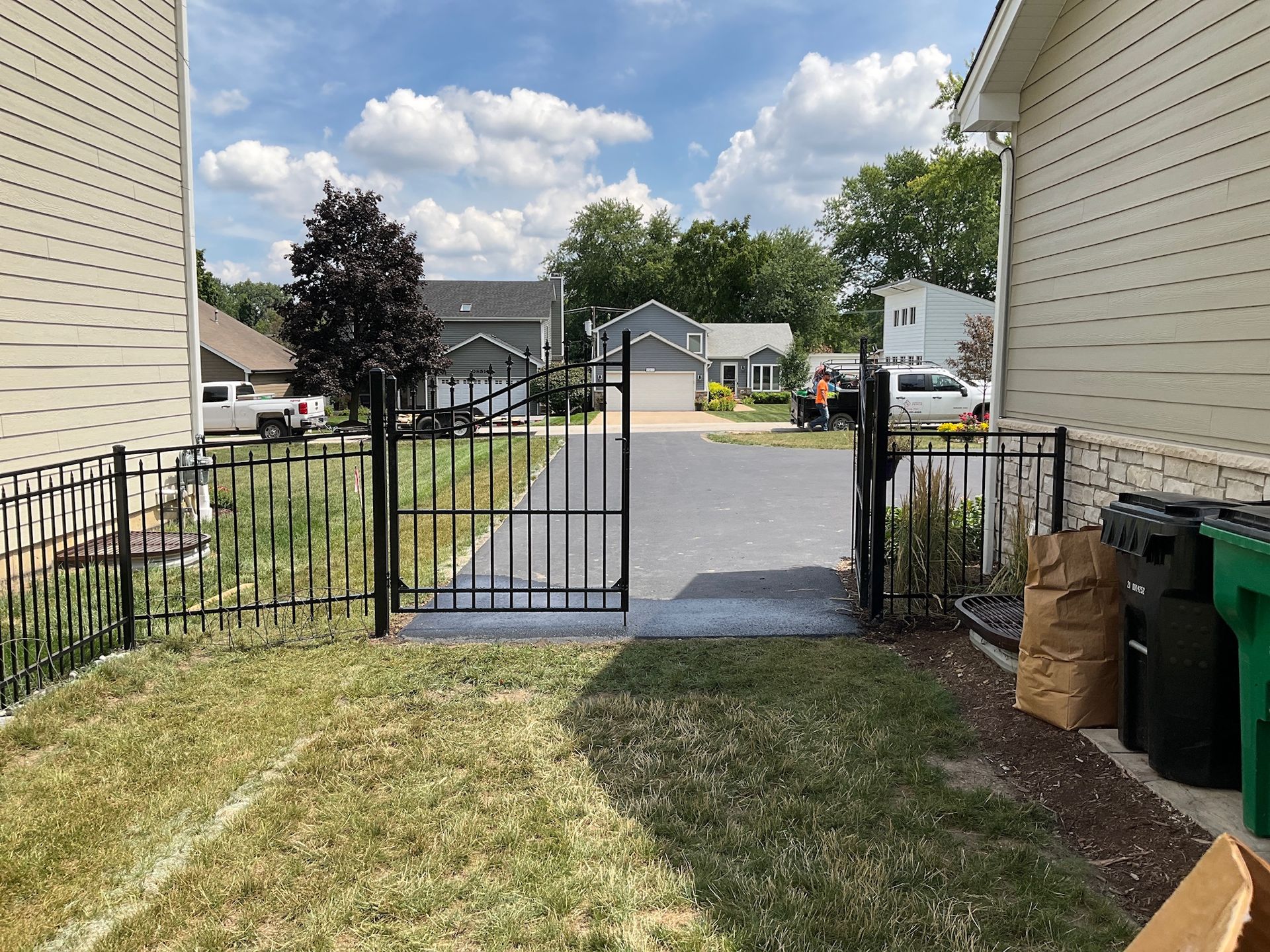 Black metal gate opens to a paved driveway between two houses, green grass foreground, sunny day.