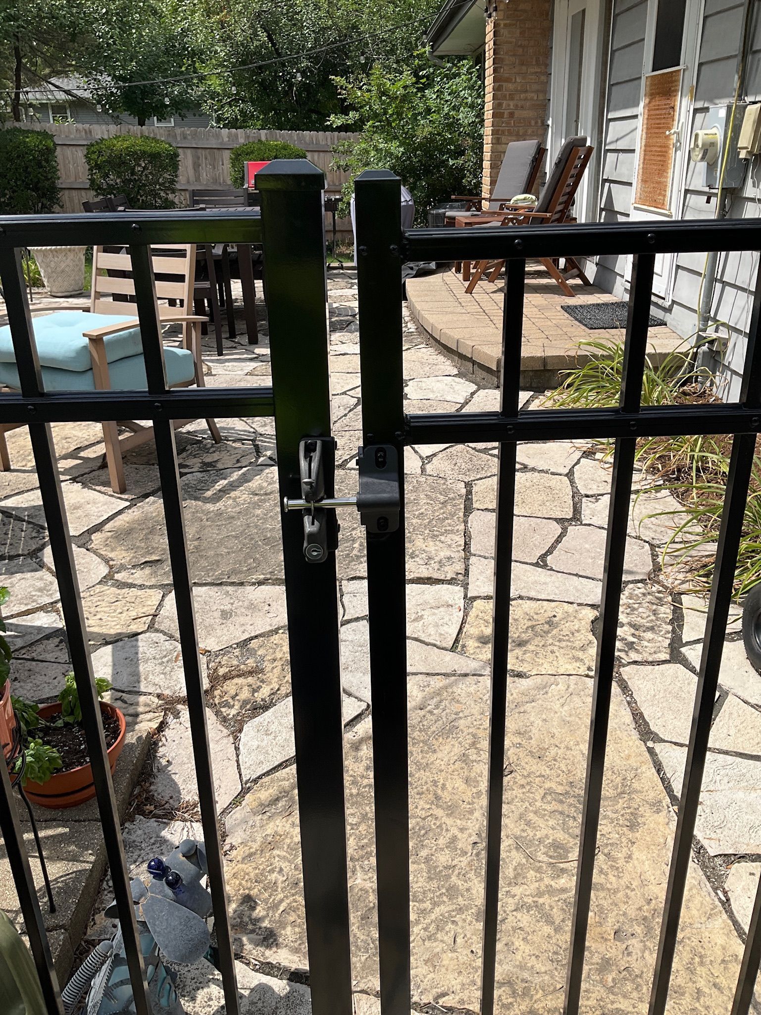 Black metal gate with latch, on a stone patio, leading to a house.