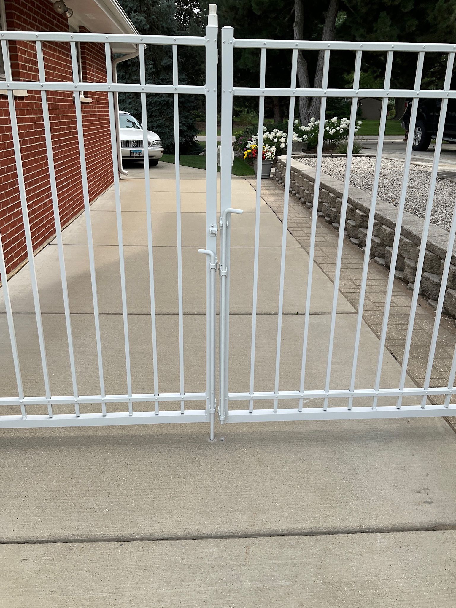 White metal driveway gates, closed, on a concrete surface, with a car visible beyond them.