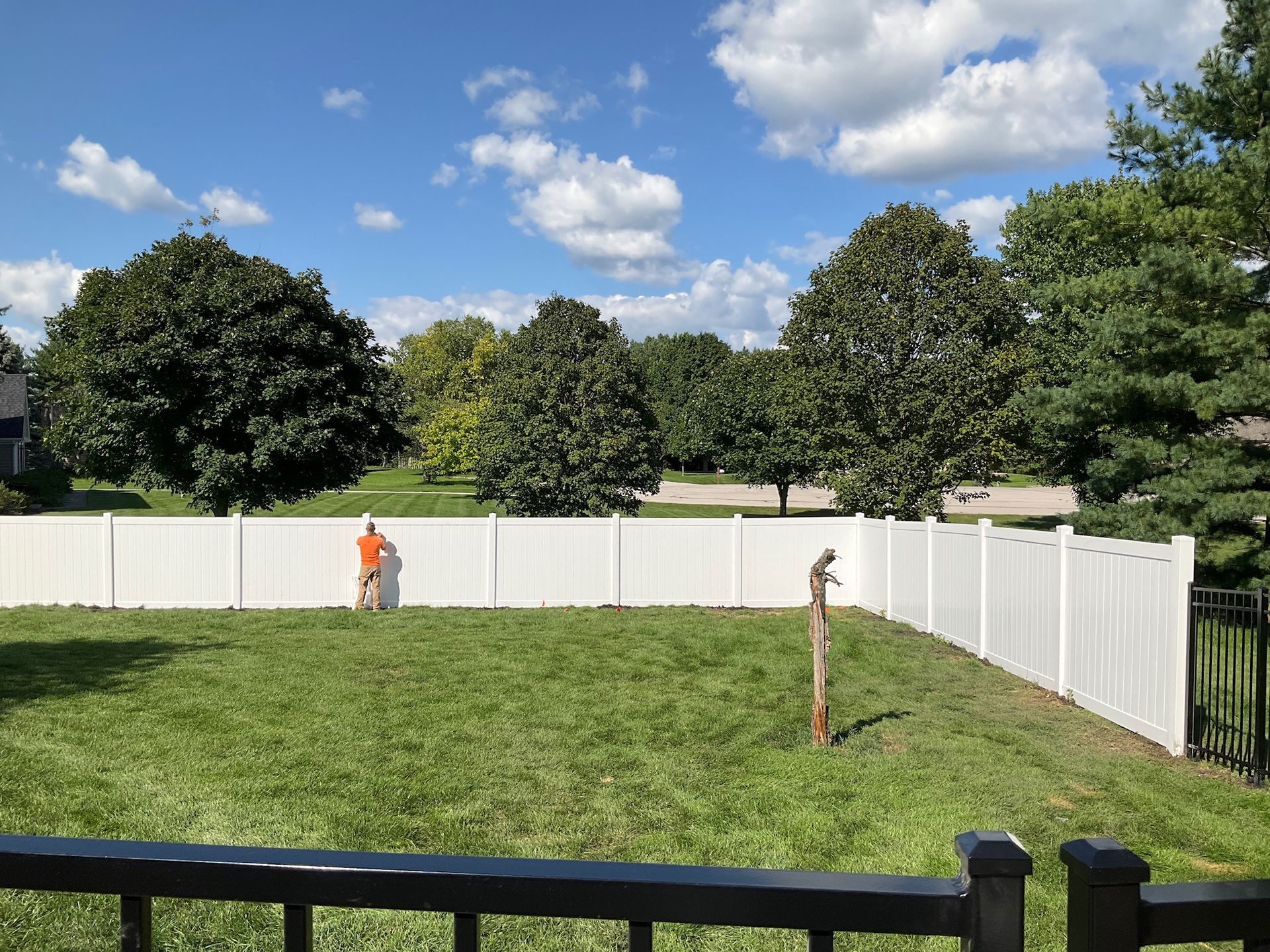 A person standing next to a white fence in a grassy backyard on a sunny day.