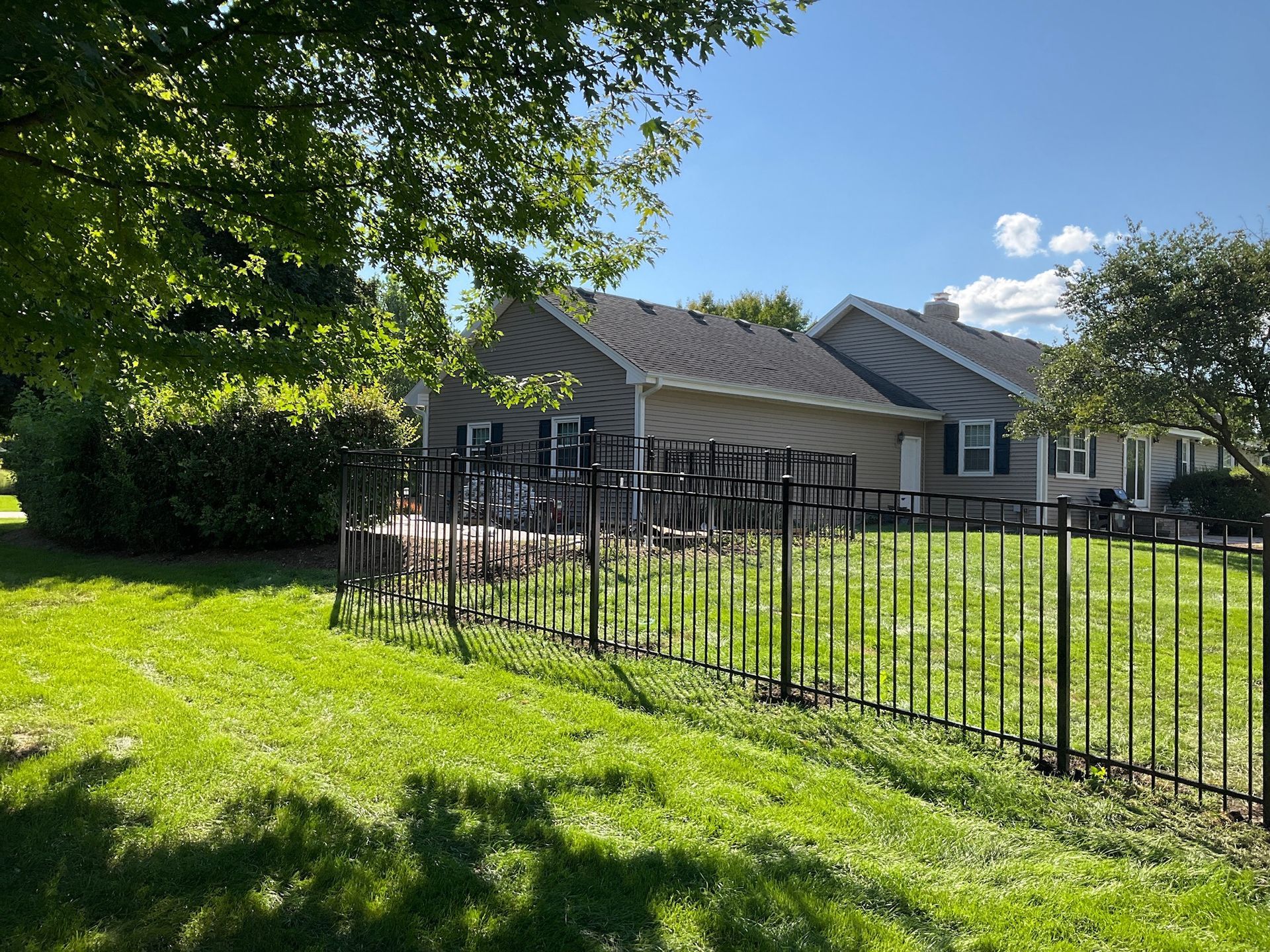 Black metal fence surrounds a grassy yard with a view of a light-colored house under a sunny sky.