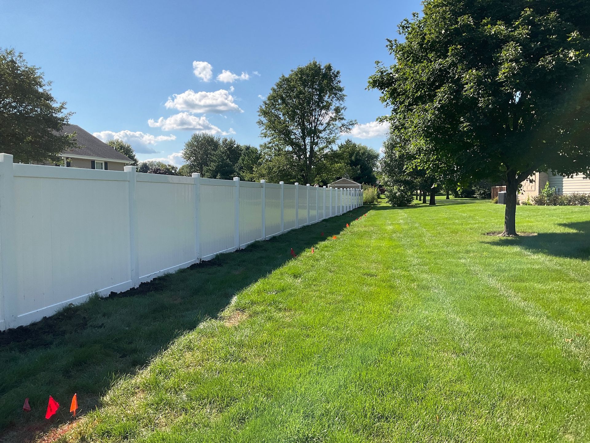 White fence lines a grassy yard on a sunny day with trees and a blue sky in the background.