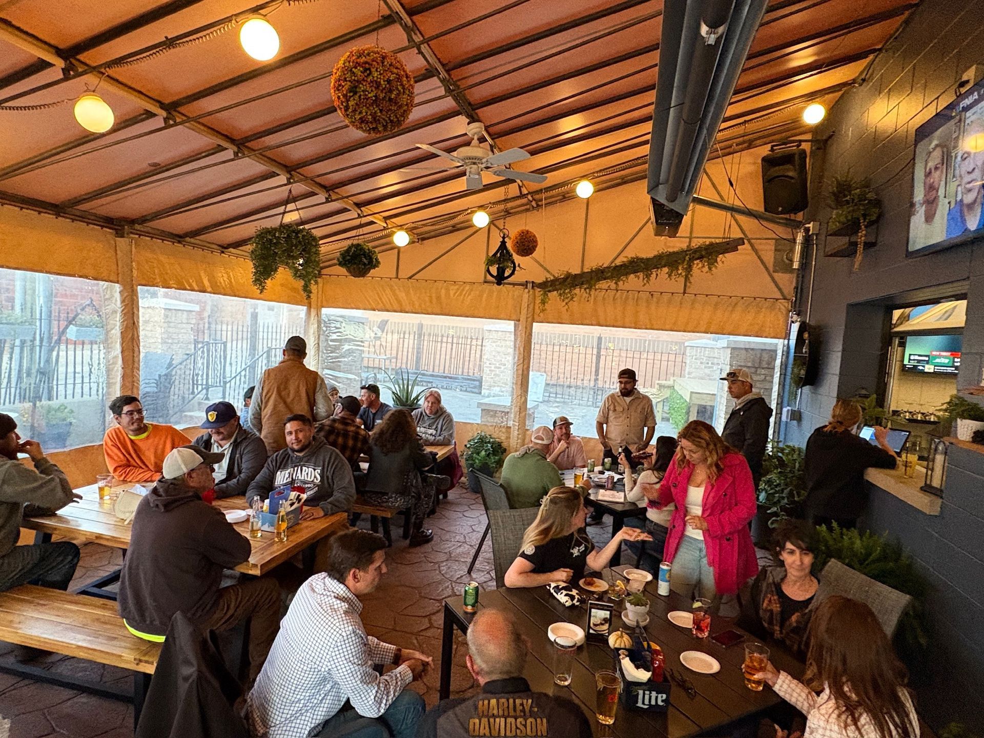 Outdoor patio with people gathered at tables, drinking and talking under a canopy.