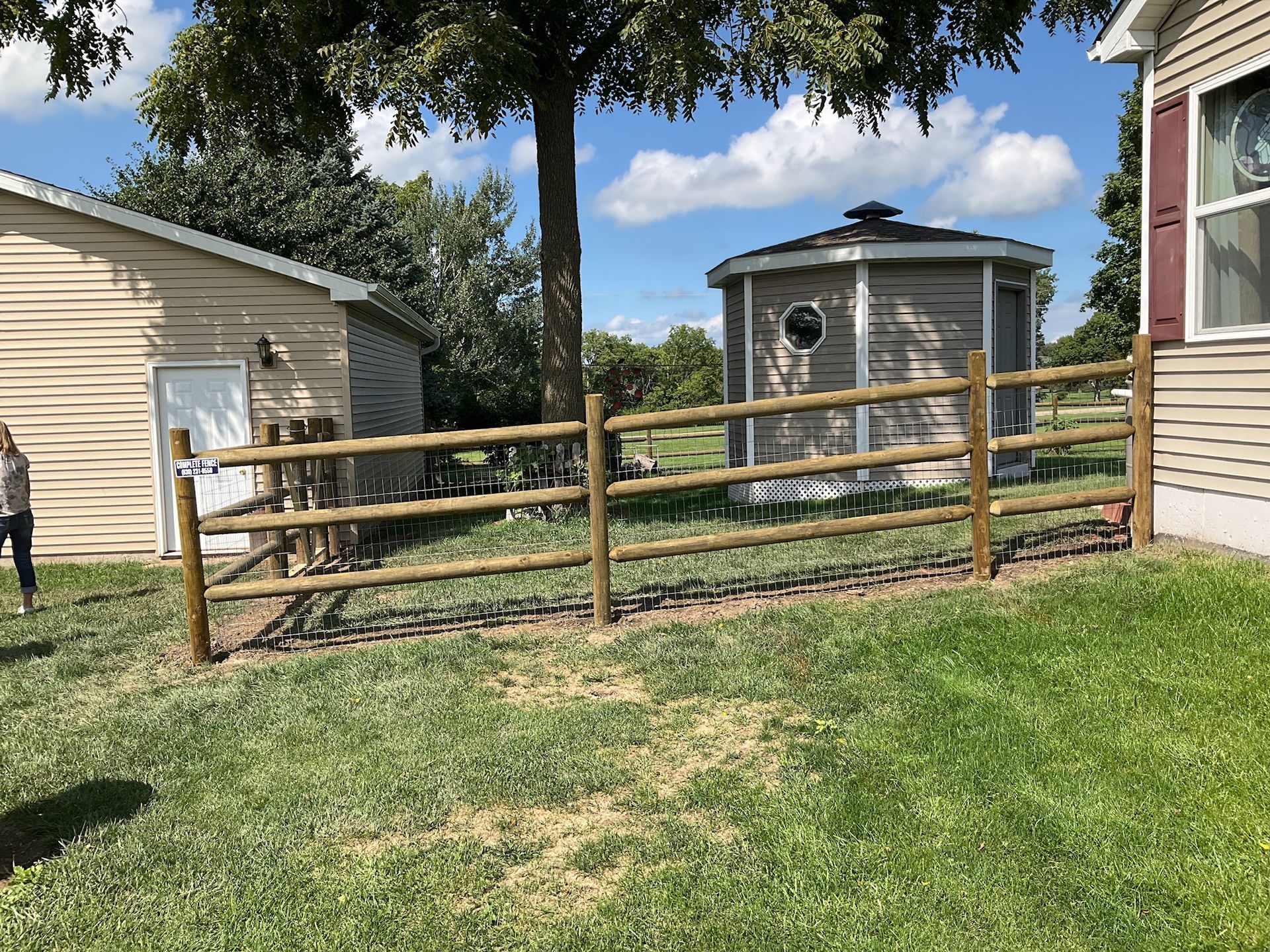 A split-rail fence surrounds a grassy area between a shed, gazebo, and house.
