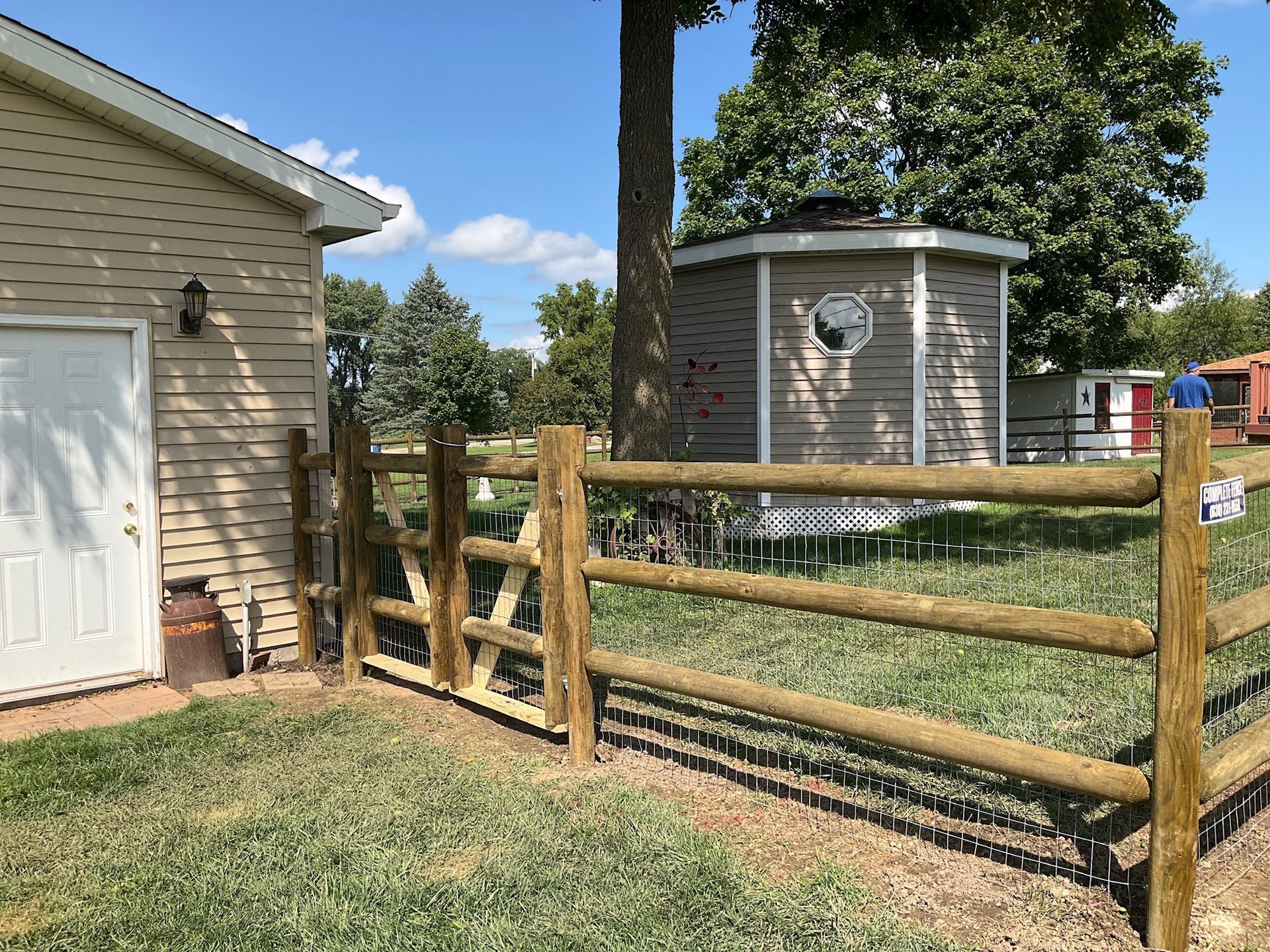 Wooden fence in a grassy yard, with a shed and house in the background on a sunny day.