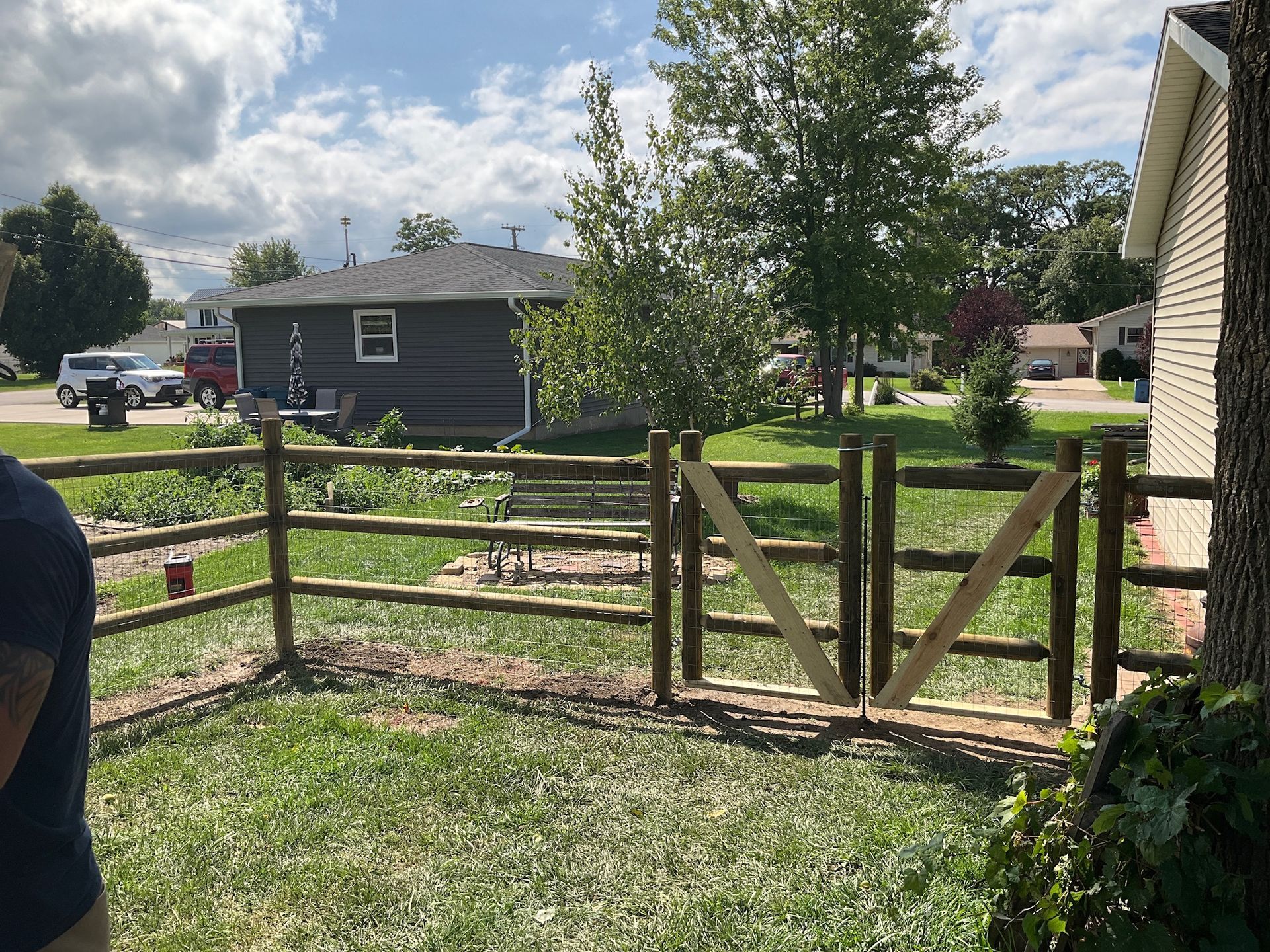 Wooden split-rail fence with gate encloses a grassy yard. A small house is in the background.
