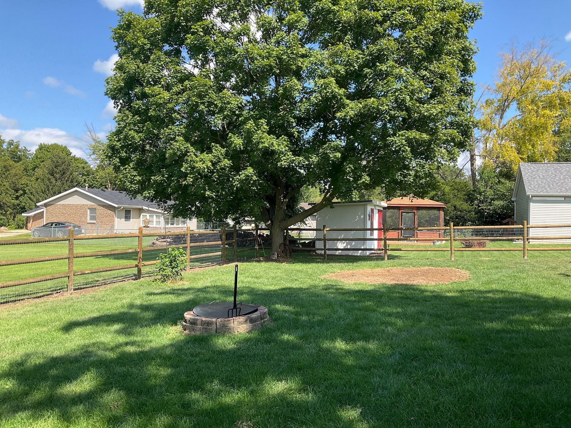 Green backyard with tree, fire pit, small shed, and wooden fence on a sunny day.