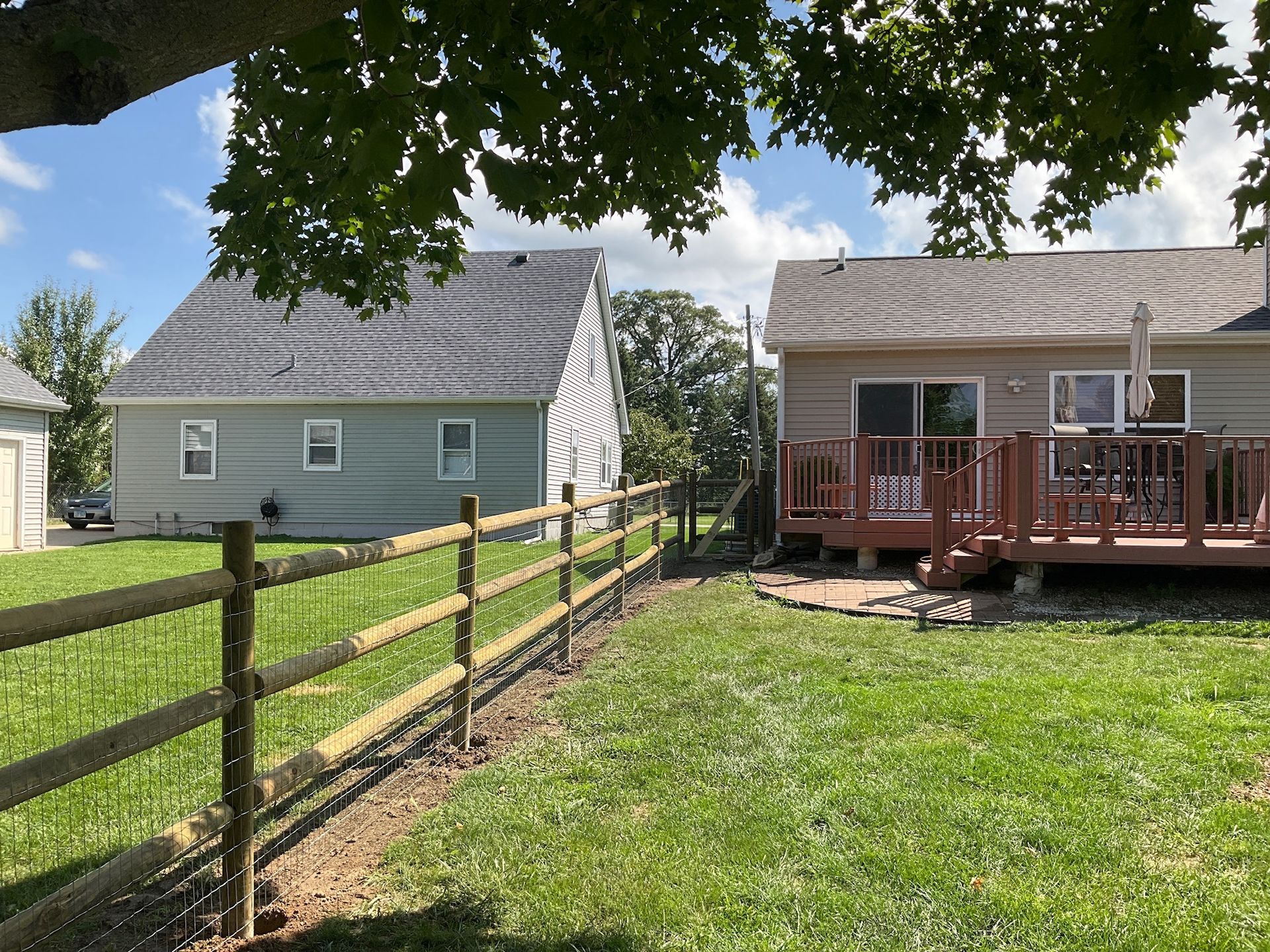 A wooden fence lines a grassy yard, separating it from two houses on a sunny day.