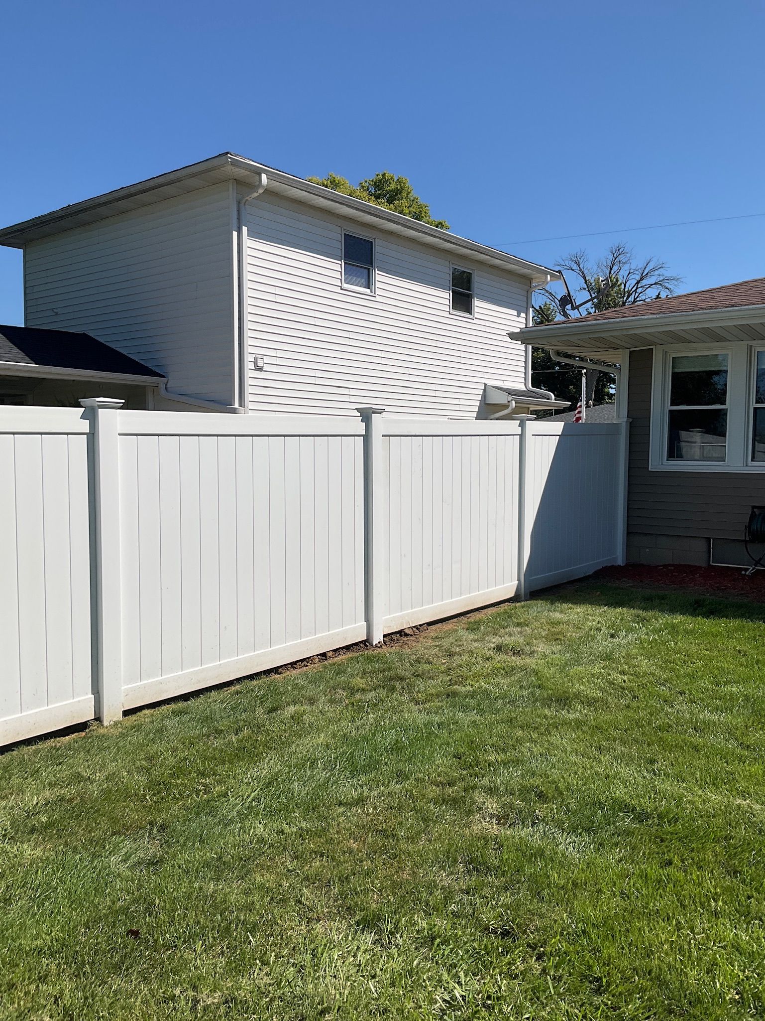 White privacy fence in a sunny backyard with two-story house visible in the background.