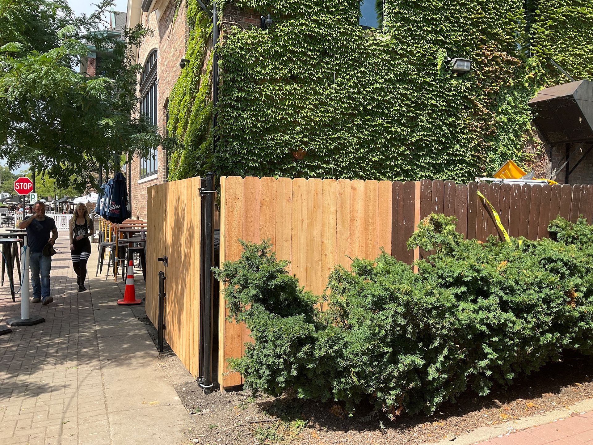 Wooden fence on a sidewalk near a building with ivy. People walk by.