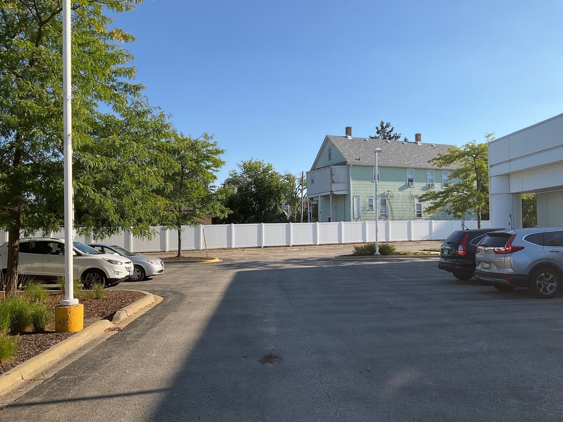 Cars parked in a paved lot; white fence borders an open area with a light green house visible in the background.