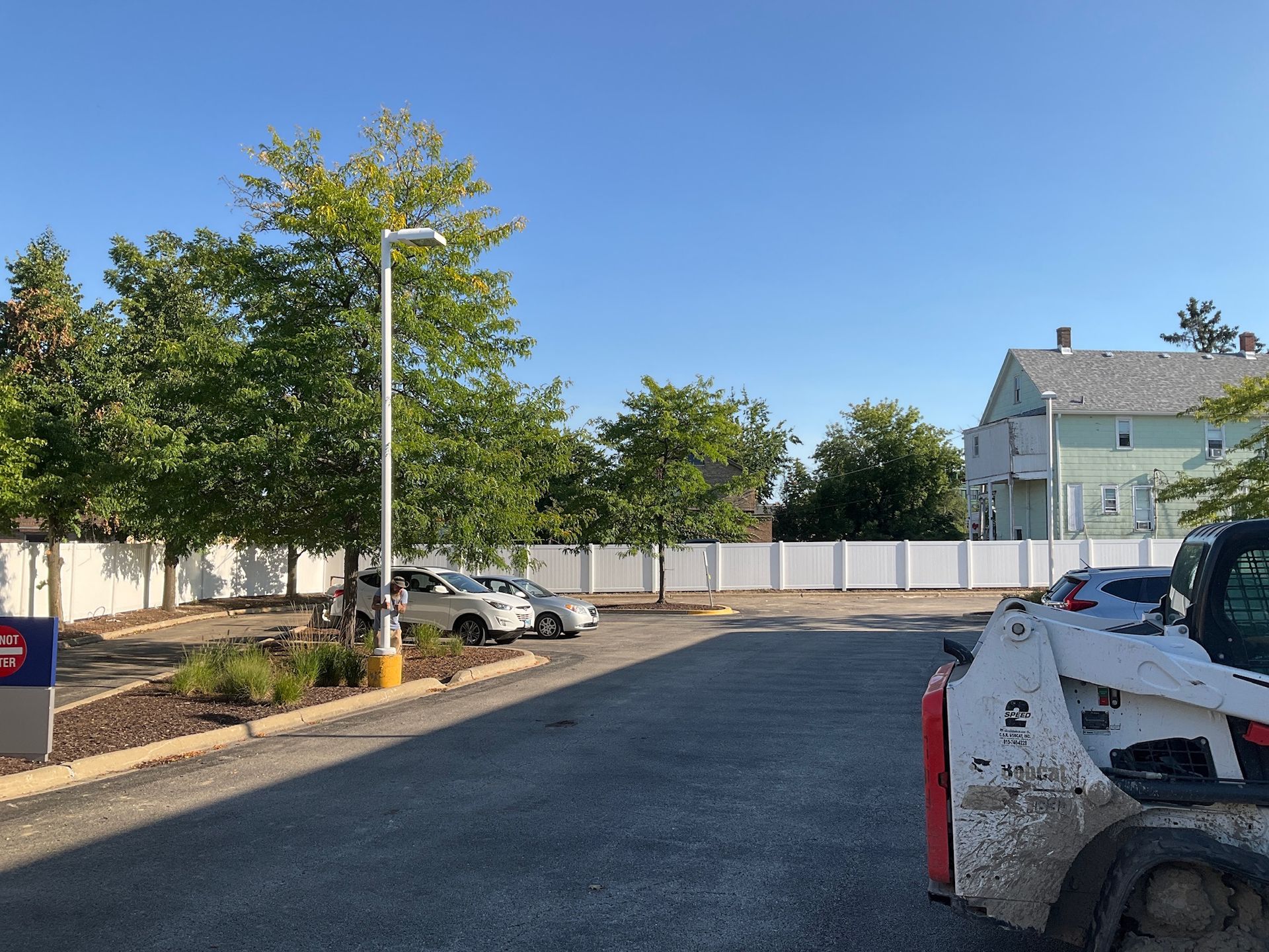 Parking lot with a white fence and trees; a house in the background.