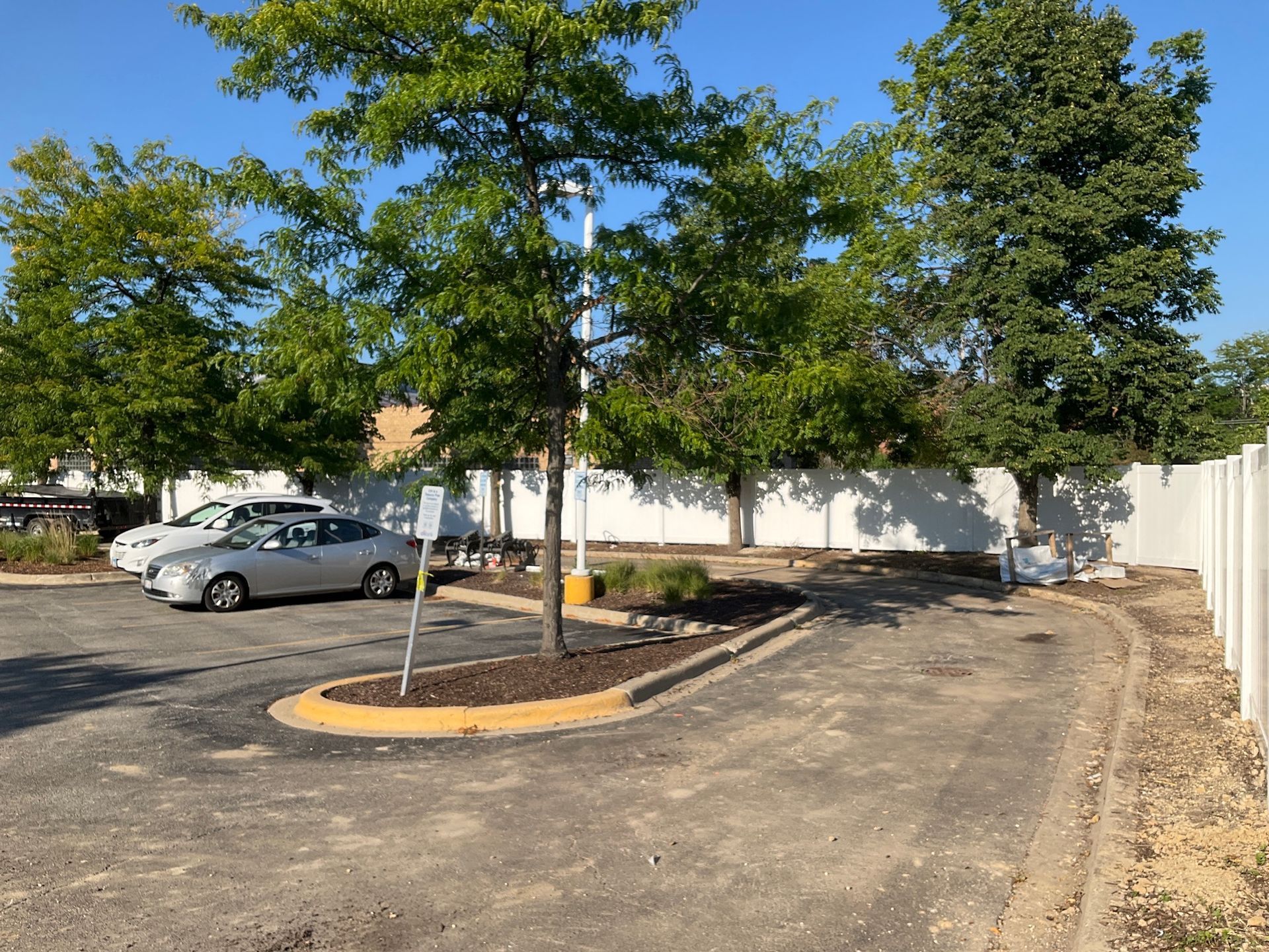 Parking lot with cars, trees, and white fence on a sunny day.
