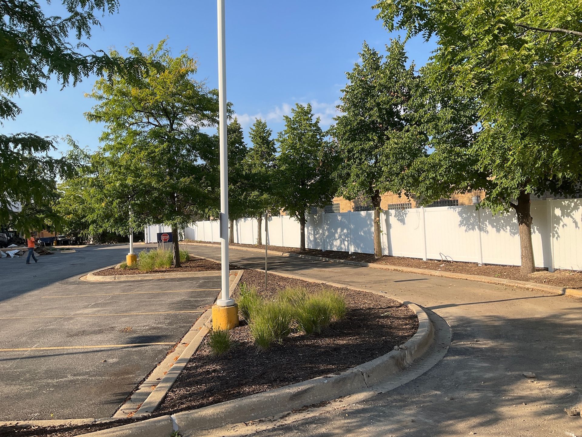Parking lot with trees and white fence under a blue sky.