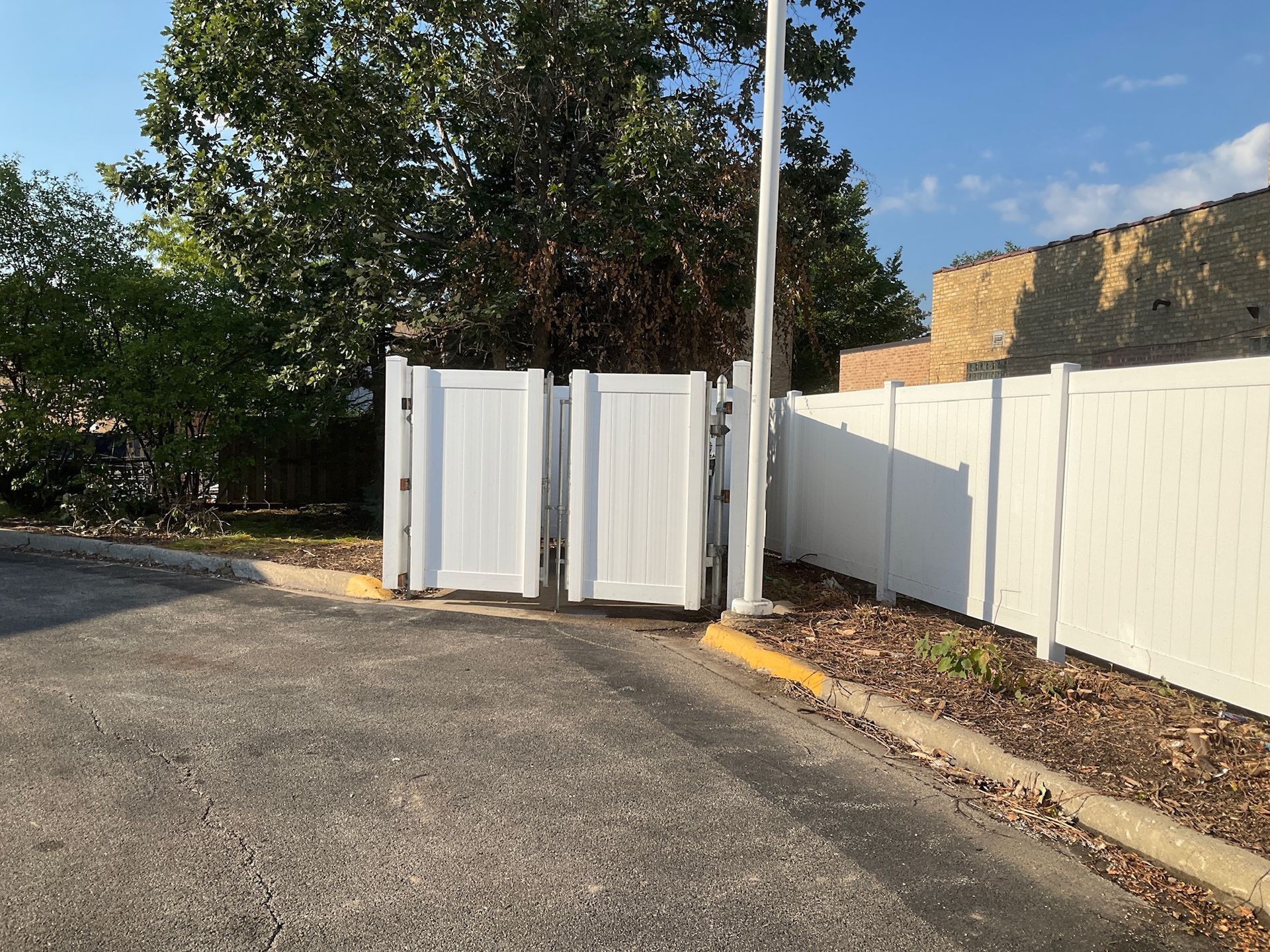 White double gates open to a parking area, flanked by white fencing and a brick building.