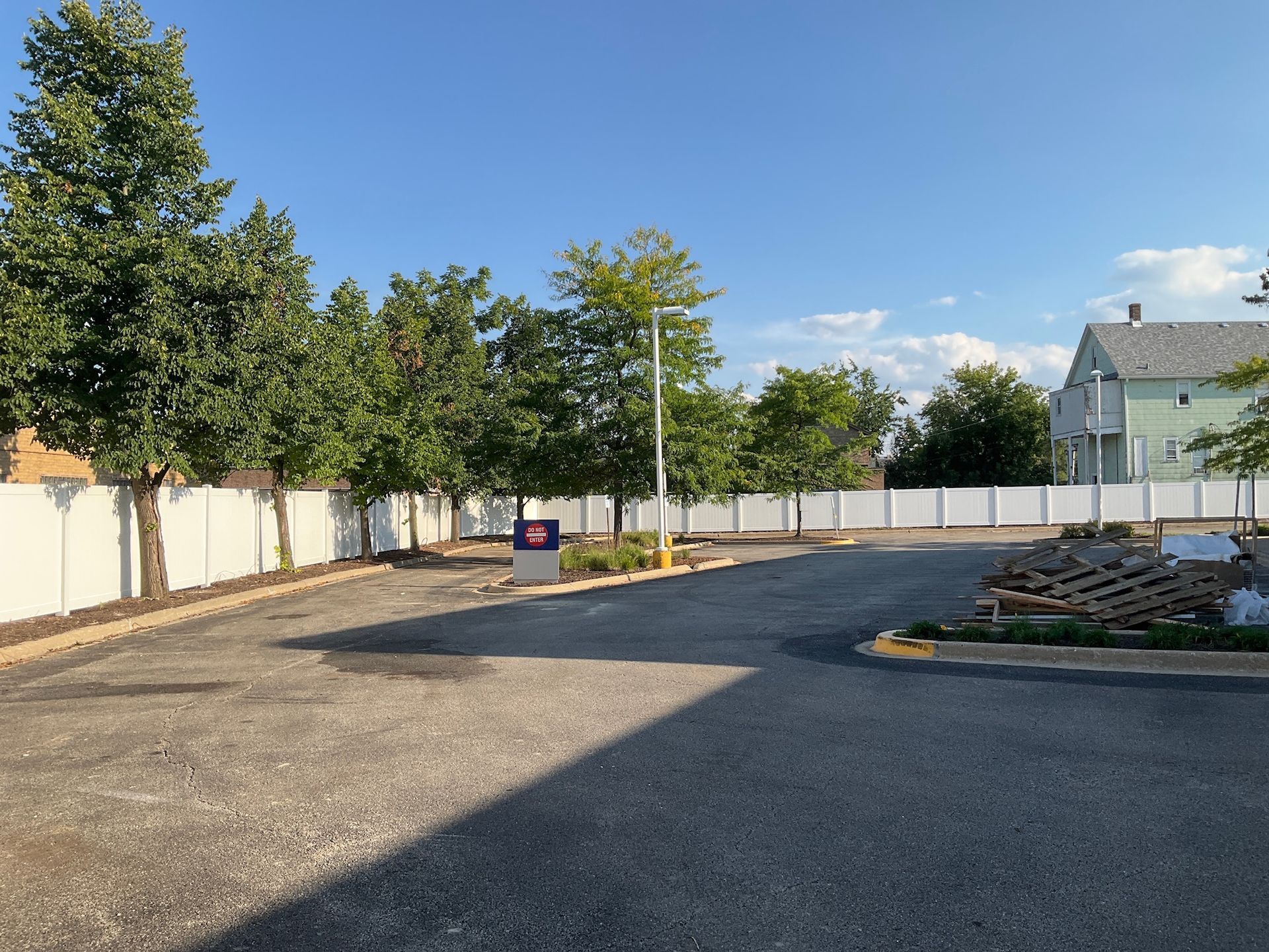 Asphalt parking lot enclosed by white fence; trees line the fence. Sunny sky.