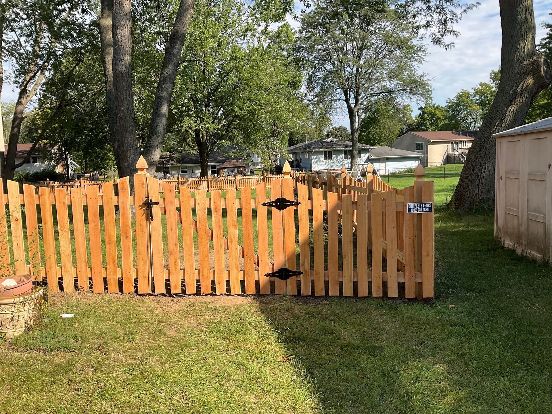Wooden picket fence and gate in a grassy yard, with trees and houses in the background.