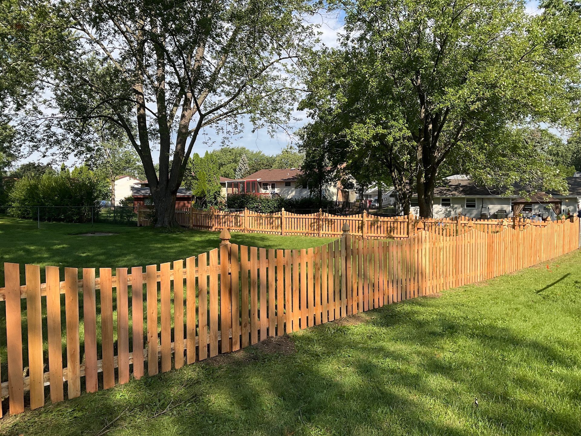 Wooden picket fence bordering a grassy yard with trees and houses in the background.