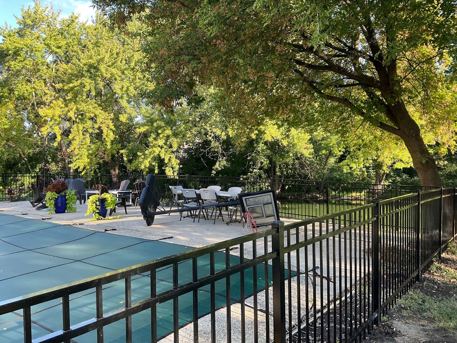 Black fence surrounds a pool with a green cover. Tables and chairs are under trees.