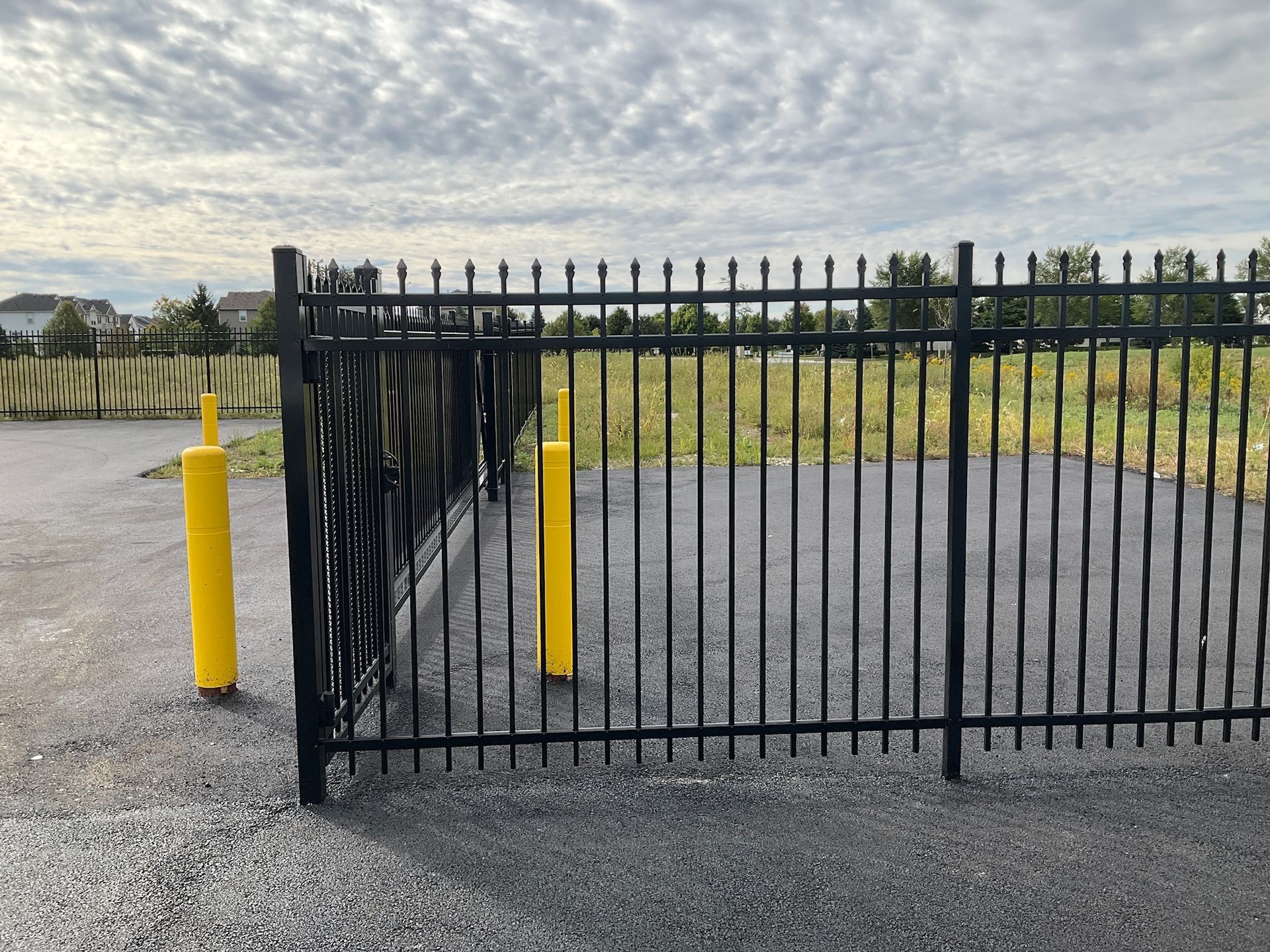 Black metal fence with pointed tops and yellow bollards on asphalt, cloudy sky background.