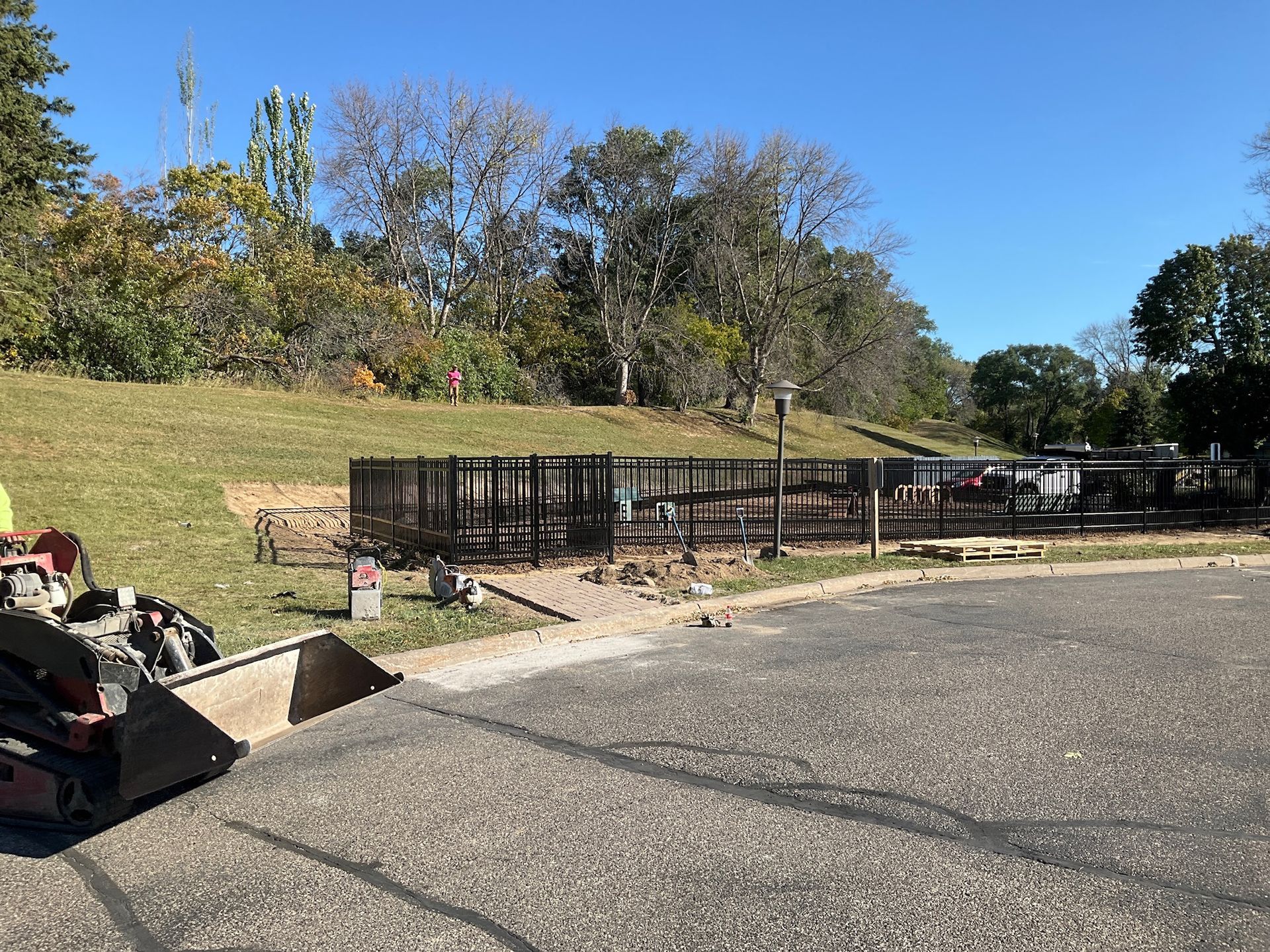 Construction site with metal fencing next to grassy hill and asphalt, small machinery in foreground.