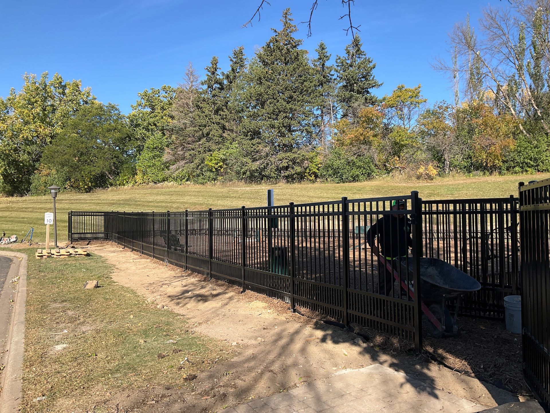 A fenced dog park with a dirt path. A person is inside the fence. Trees and a blue sky are in the background.