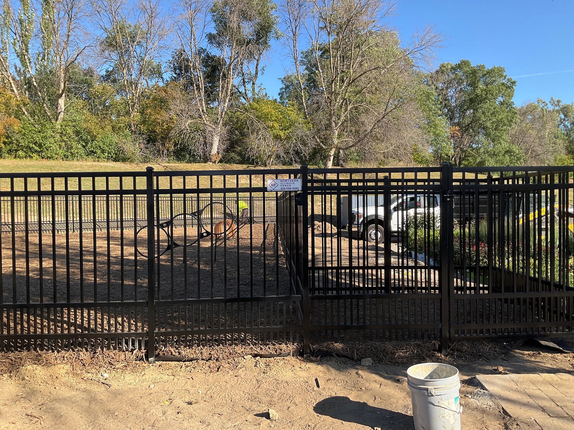 Black metal fence surrounding an enclosure with a gate, trees in the background, and a white bucket in the foreground.