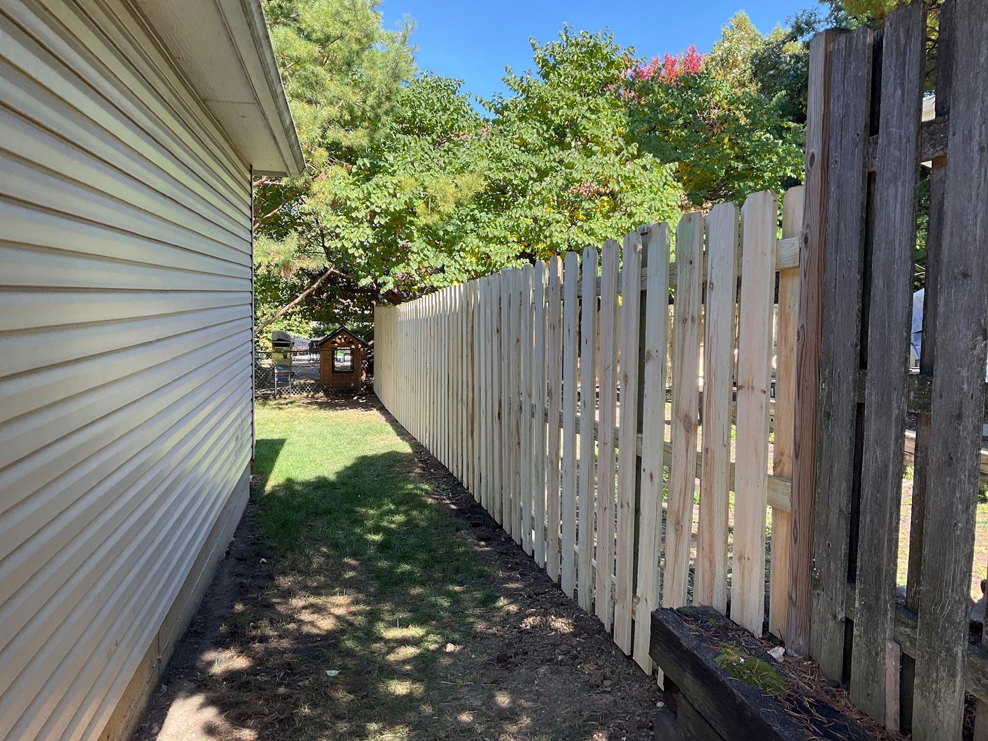 Side of a house and weathered wooden fence, with green grass in between, under a sunny sky.