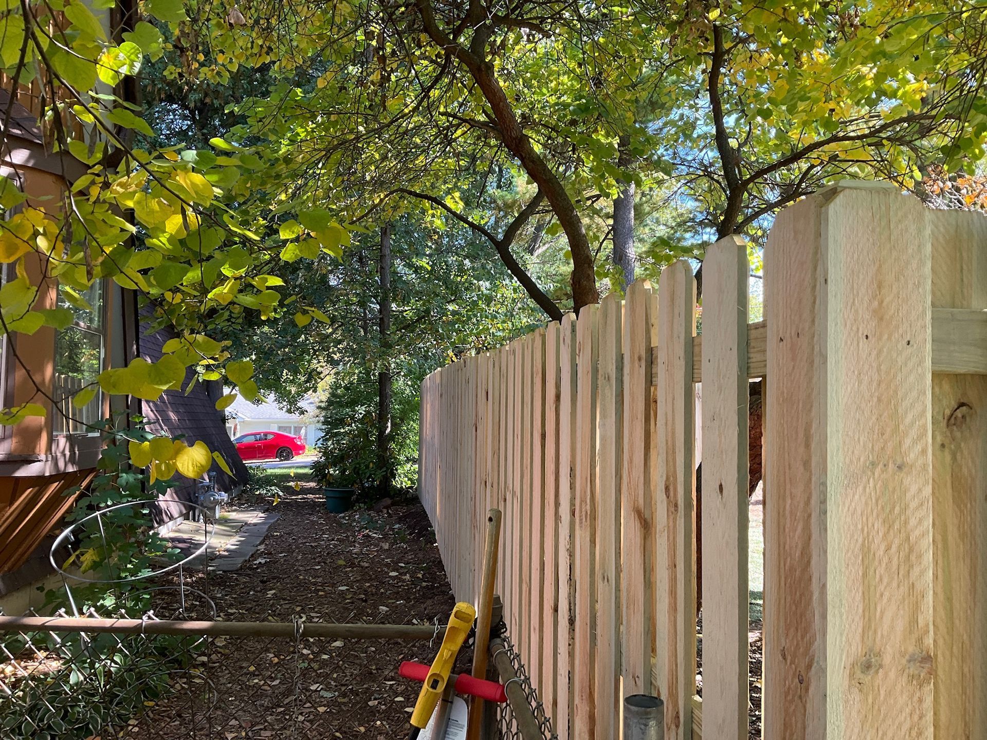 Wooden fence in a yard with leaves, a red car, and a tree in the background.