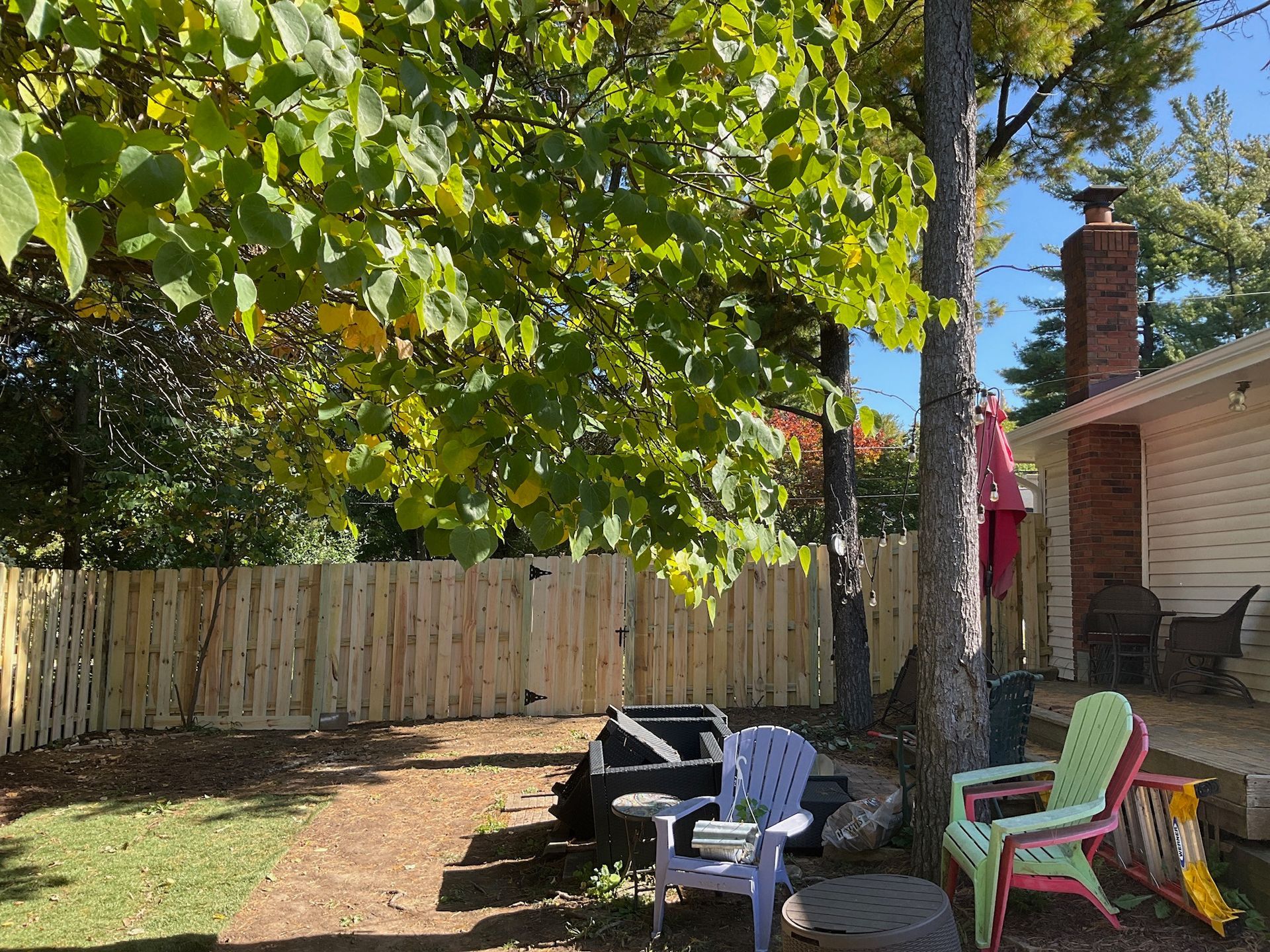 Wooden fence encloses a backyard with trees, chairs, and a chimney.