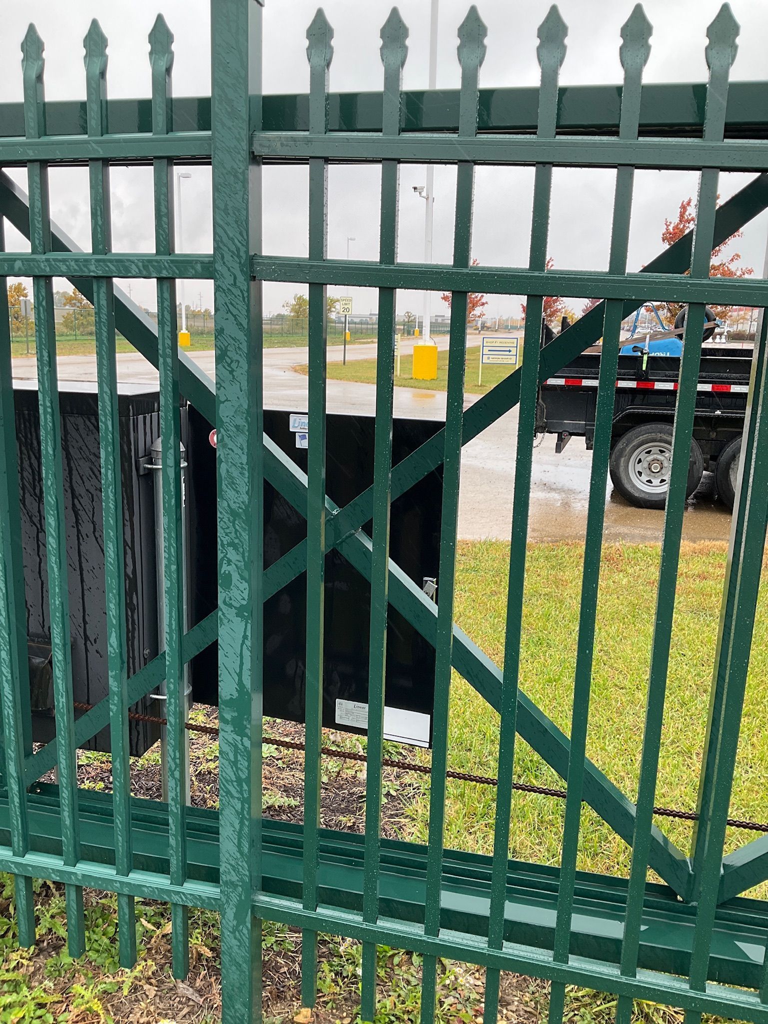 Green metal fence with a black trash enclosure in the background, a truck is partially visible.