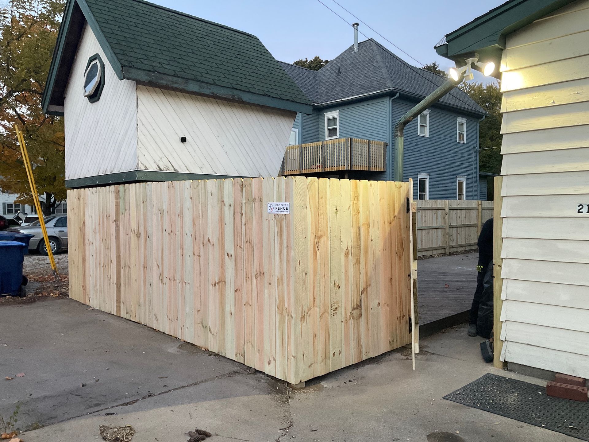 Wooden fence encloses an outdoor area, with buildings in the background and a paved surface in front.