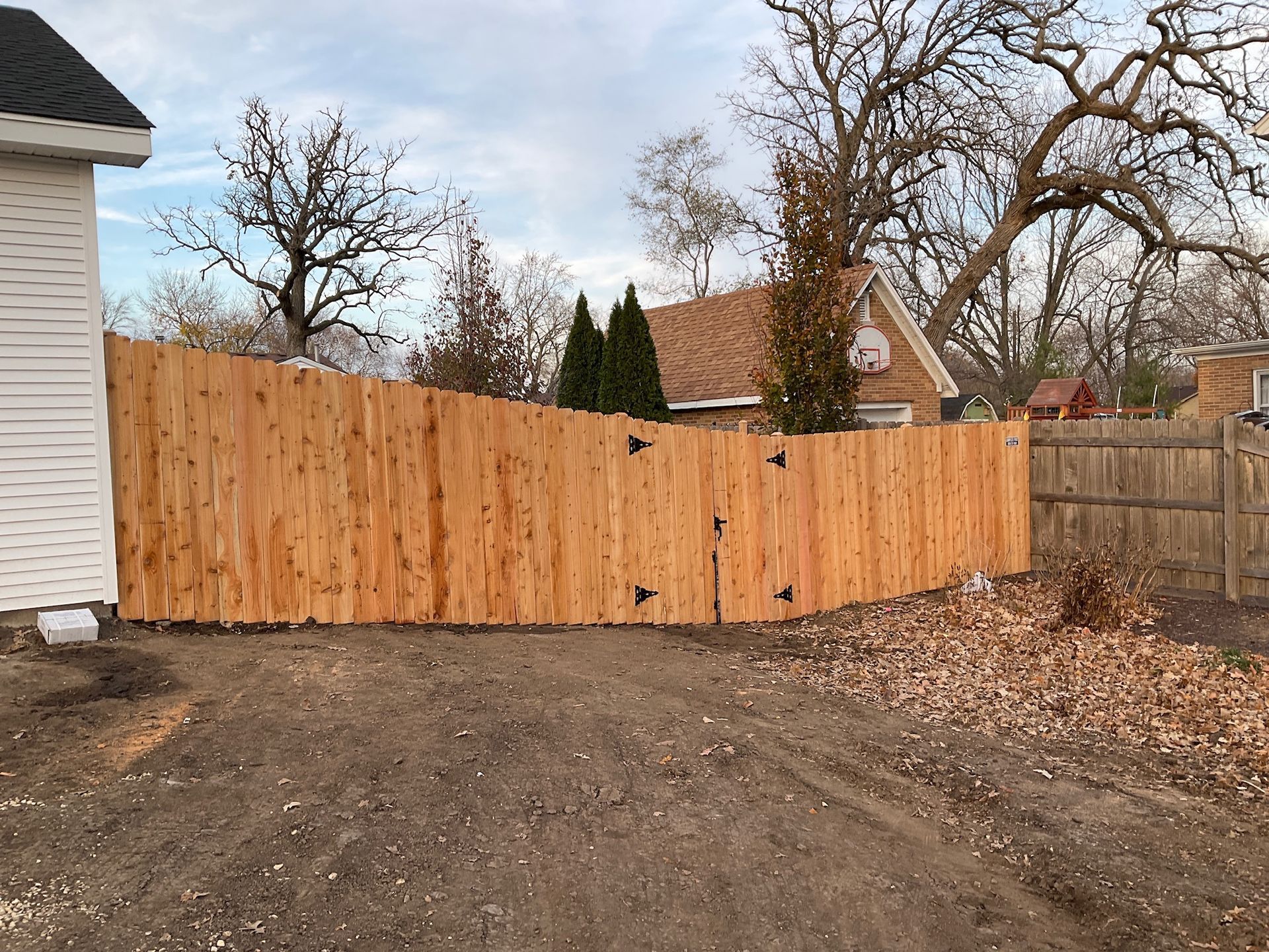 Wooden privacy fence in a yard, gate with black hinges, trees in the background, overcast sky.