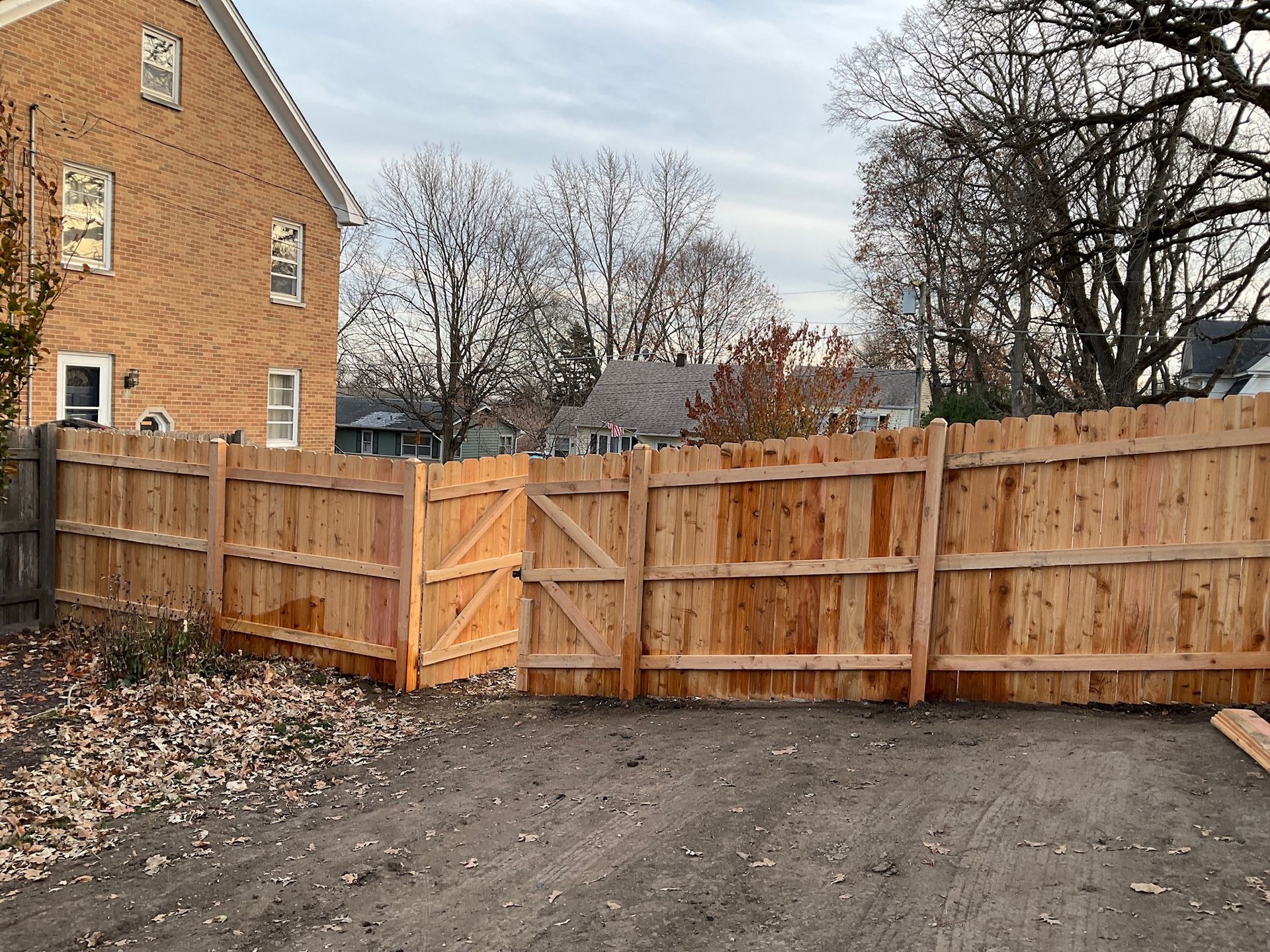 Wooden fence with gate in front of a brick building and trees under a cloudy sky.